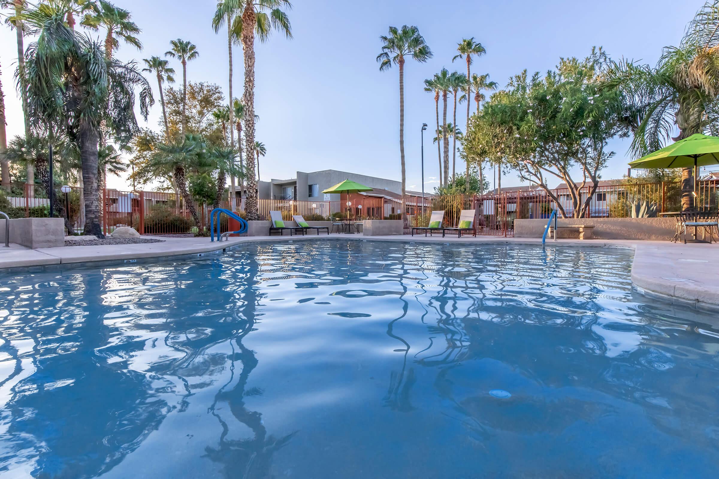 A clear swimming pool surrounded by palm trees and lounge chairs. Green umbrellas provide shade on the pool deck, with a building in the background. The setting is tranquil, showcasing a sunny sky and a relaxing atmosphere ideal for leisure and recreation.