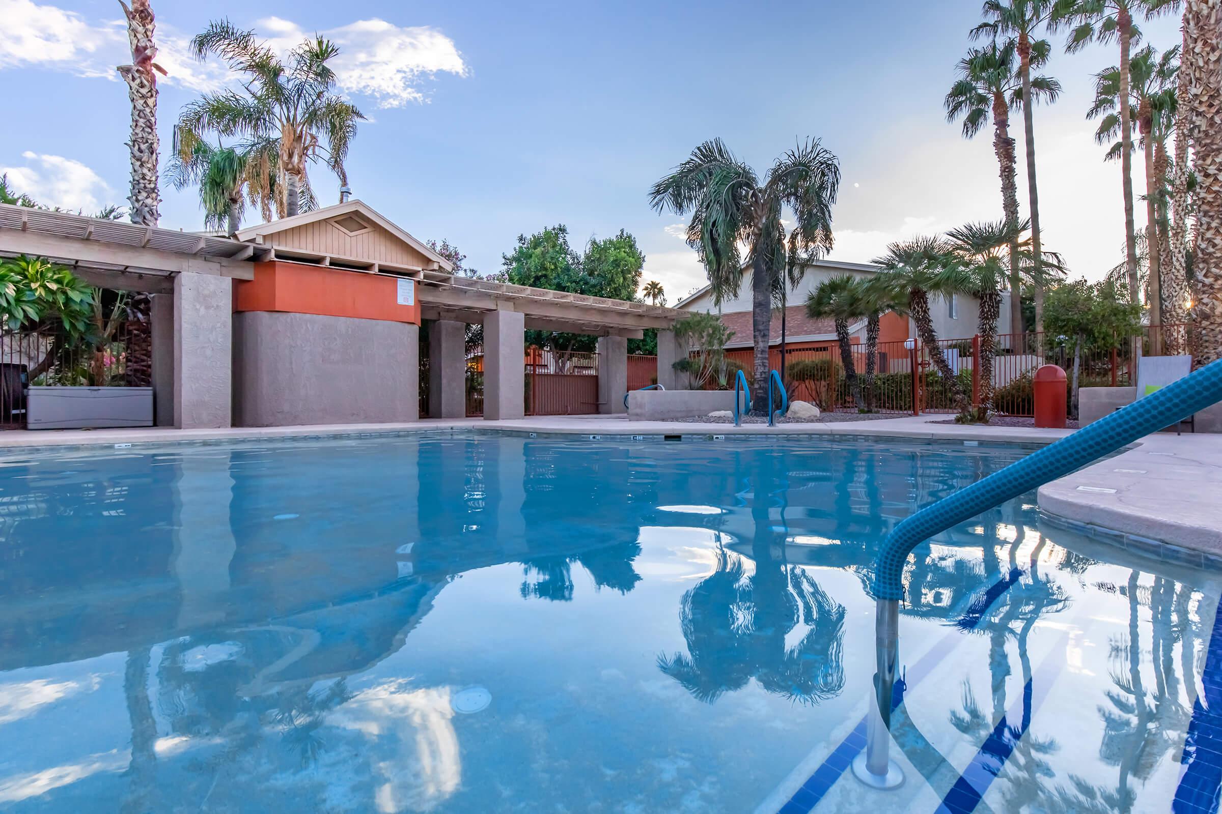 A serene swimming pool surrounded by palm trees and landscape features. The water is calm and reflecting the sky, with an inviting staircase leading into the pool. Nearby, there are seating areas and a backdrop of residential buildings. The setting suggests a tranquil outdoor retreat.