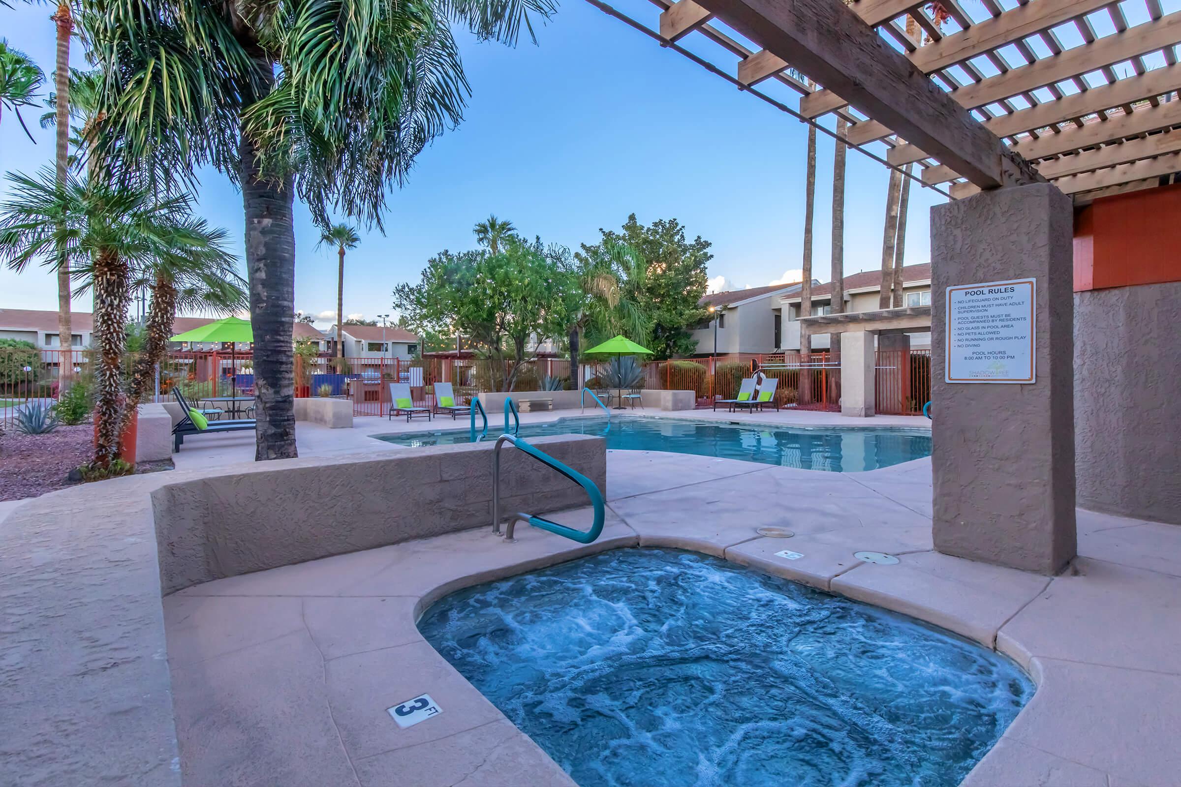 A sunny outdoor pool area featuring a large swimming pool and a hot tub. Surrounding palm trees and lounge chairs create a relaxing atmosphere. A shaded area with umbrellas can be seen in the background, along with a sign displaying pool rules. The scene is bright and inviting, ideal for leisure and relaxation.