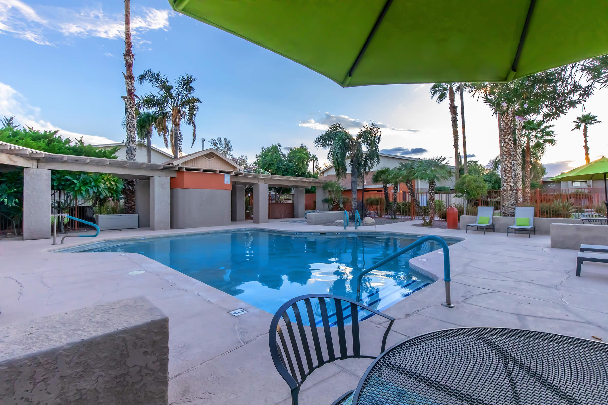 A view of a swimming pool surrounded by palm trees and green umbrellas. The patio features a table and chairs overlooking the pool area, which has lounge chairs and a tranquil atmosphere. The sky is partly cloudy, and the setting suggests a relaxing outdoor environment.