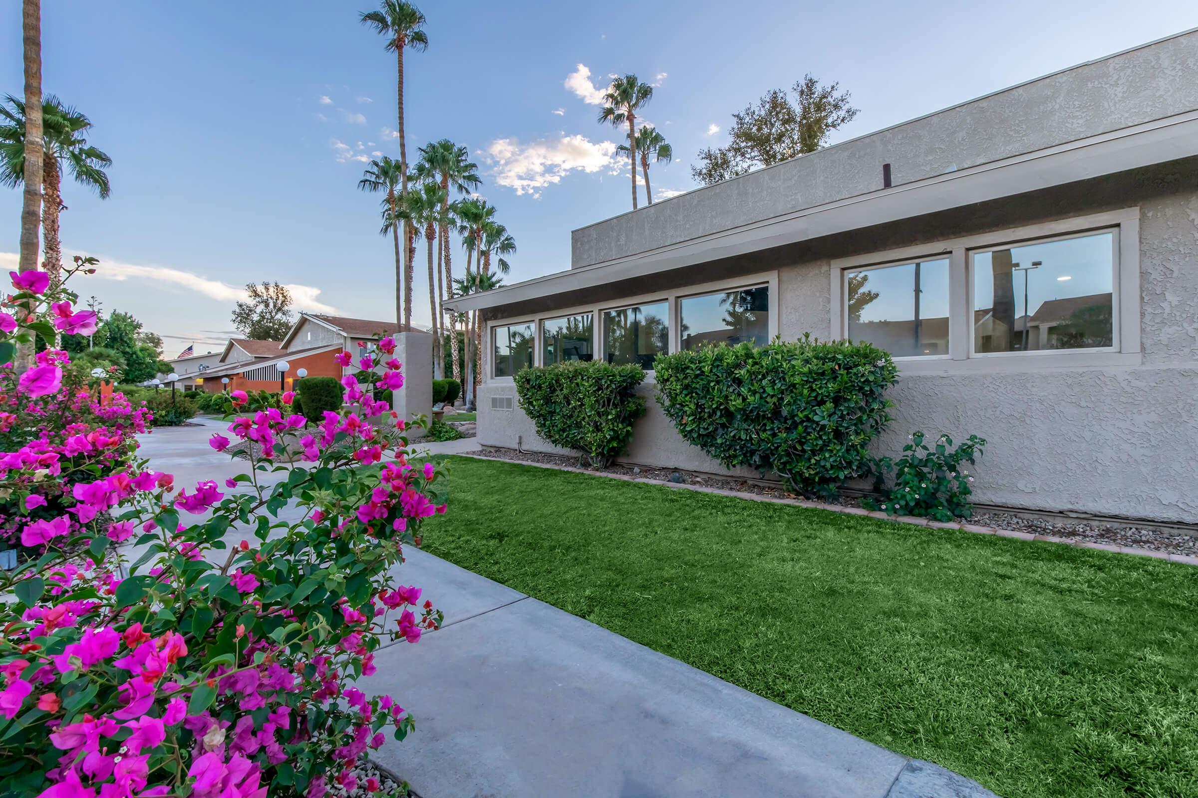 A landscaped exterior view of a building featuring a pathway lined with vibrant pink flowers and manicured grass. Tall palm trees are visible in the background under a clear sky. The setting conveys a warm, inviting atmosphere typical of a suburban area.