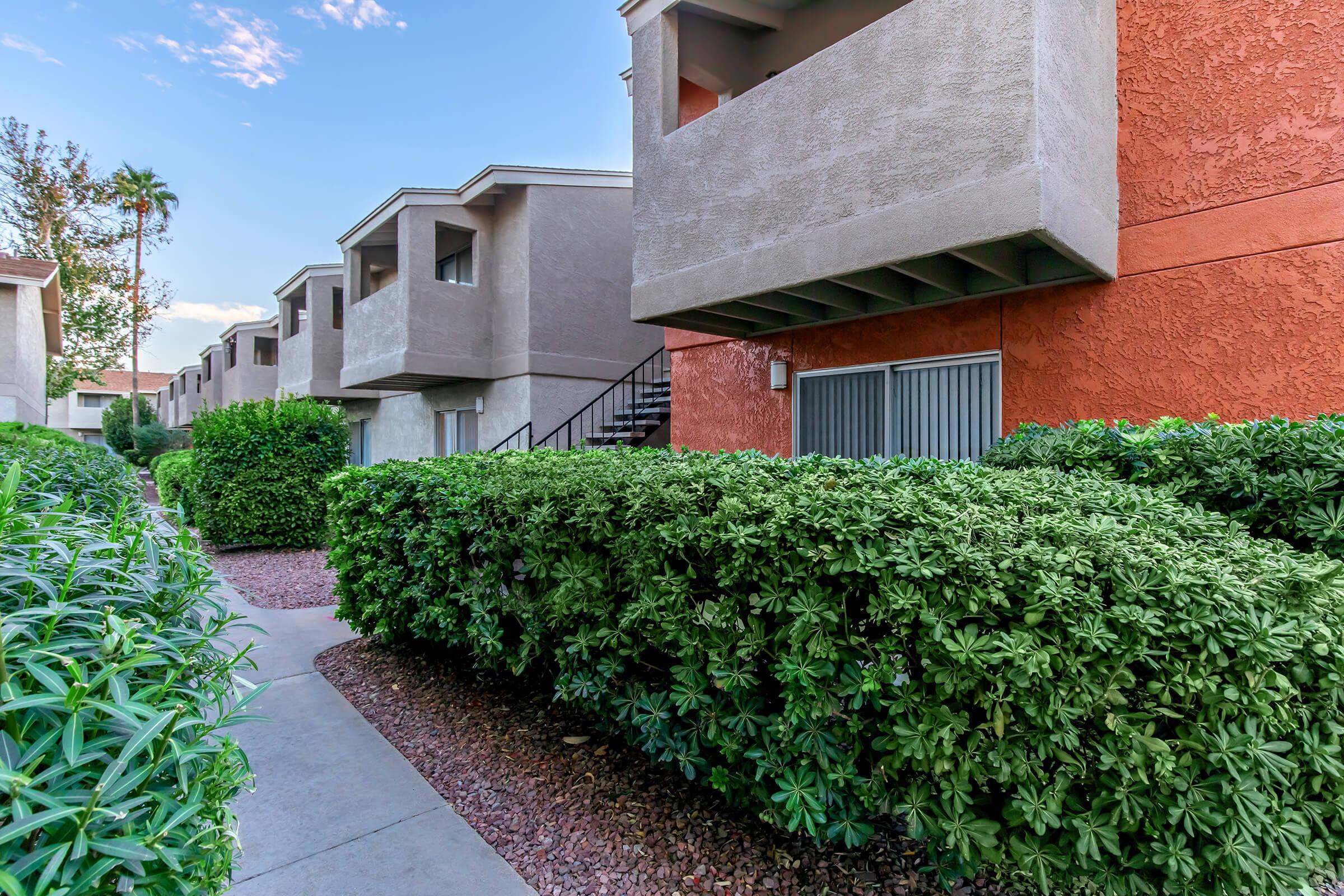 A walkway lined with lush green shrubs leads to a set of multi-story apartment buildings. The buildings have a mix of colors, including beige and orange, and features a staircase in the foreground. The scene is set against a clear sky with a few clouds.