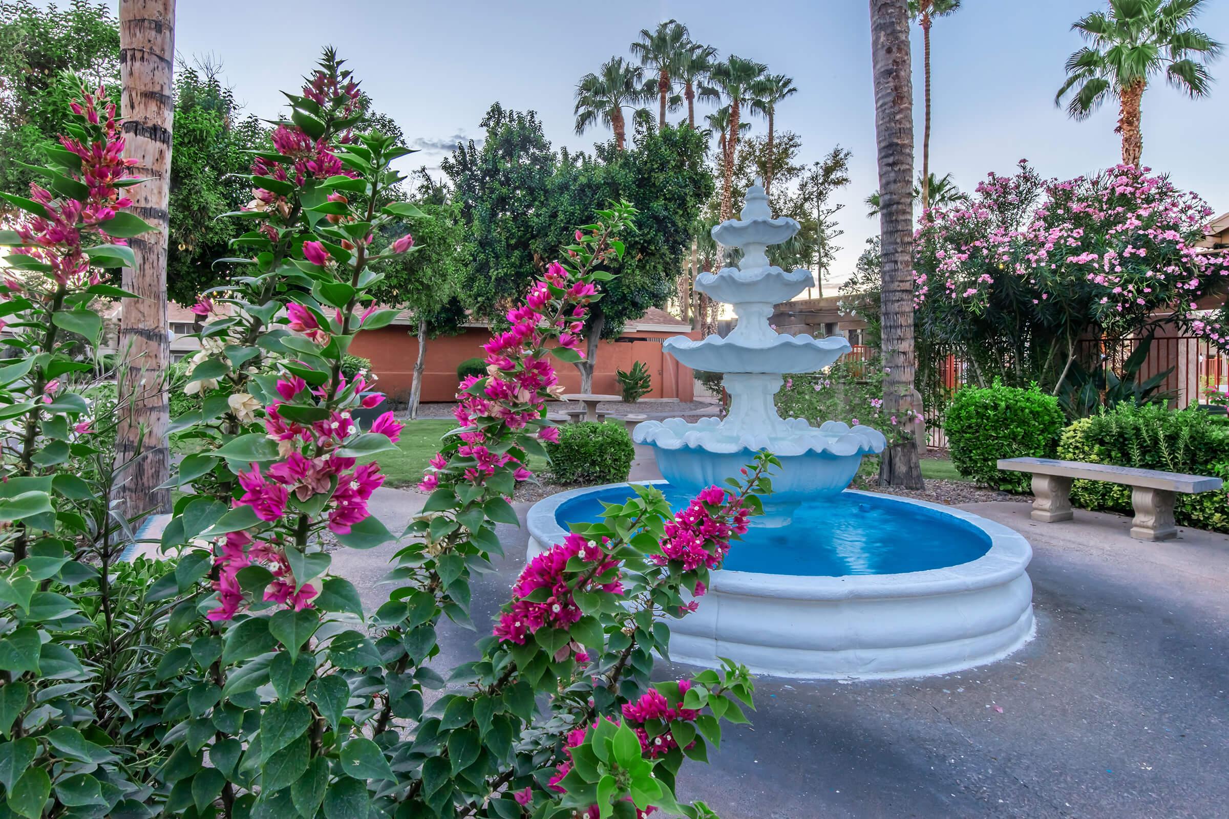 A serene garden scene featuring a blue fountain surrounded by lush greenery and vibrant pink bougainvillea flowers. Palm trees and flowering plants enhance the tranquil atmosphere, creating a peaceful outdoor space for relaxation.