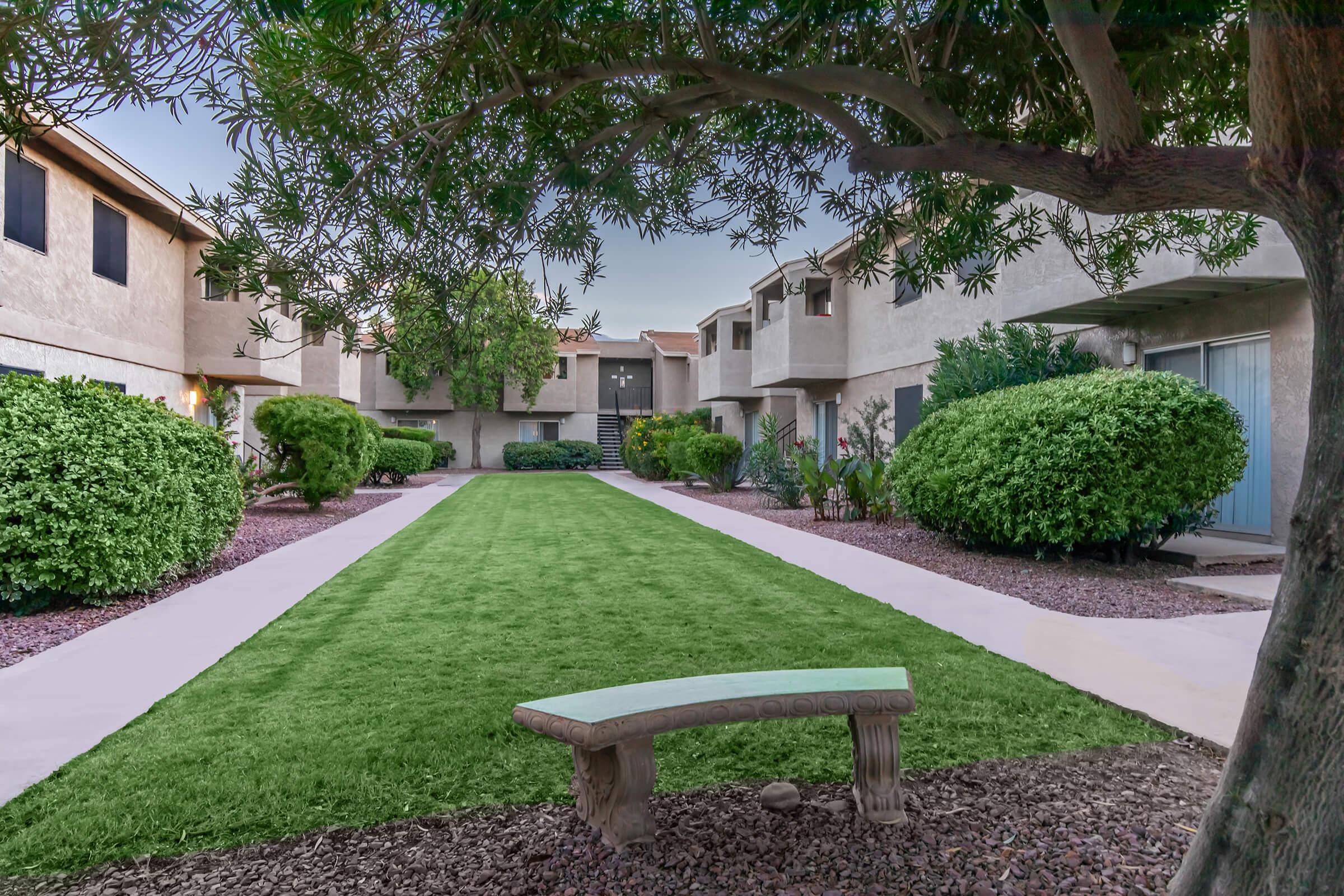 A tranquil courtyard scene featuring neatly manicured grass and hedges, flanked by residential buildings on either side. A stone bench sits along a paved pathway, inviting relaxation amidst the greenery. The atmosphere is calm and serene, perfect for enjoying outdoor moments.