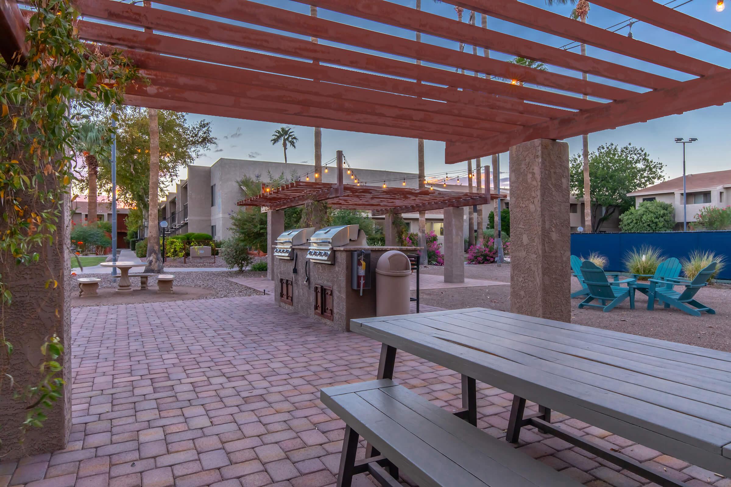 Outdoor communal area featuring a barbecue grill under a slatted pergola, with picnic tables set on a brick patio. Surrounding greenery includes shrubs and desert plants, with a glimpse of nearby buildings and palm trees in the background. Ideal for social gatherings and relaxation.