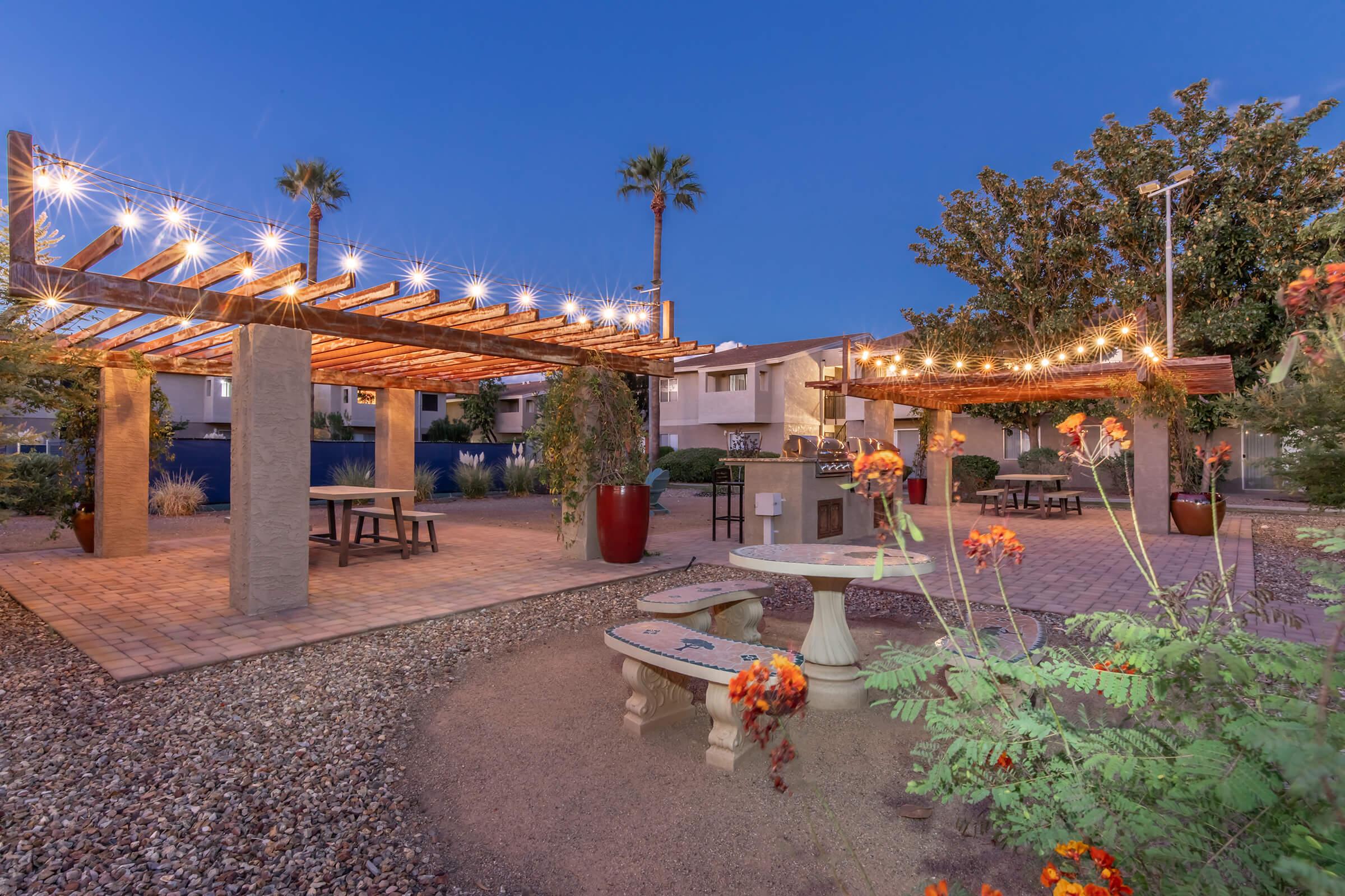 A landscaped outdoor area at dusk featuring a wooden pergola adorned with string lights, a picnic table, stone seating, and vibrant flower beds. The setting includes palm trees and residential buildings in the background, creating a cozy and inviting atmosphere for gatherings.