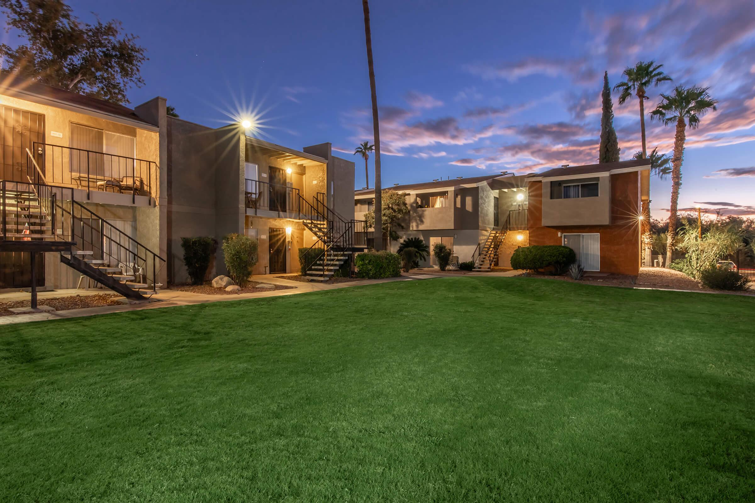 A view of an apartment complex at twilight, featuring two-story buildings with staircases, surrounded by well-manicured green lawns and palm trees. The sky showcases a colorful sunset with scattered clouds, enhancing the serene atmosphere of the residential area.