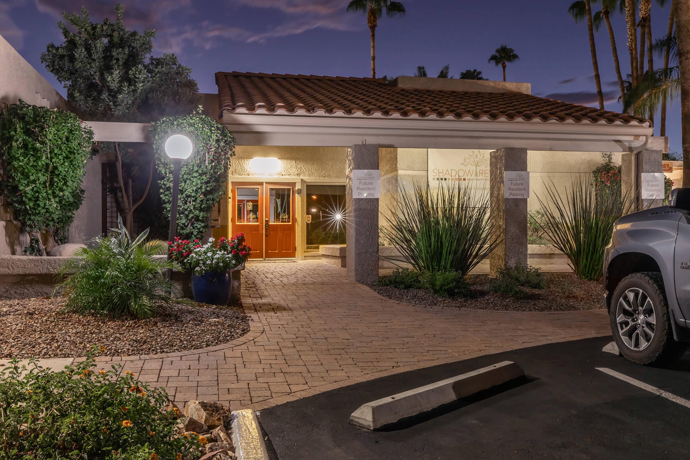 Exterior view of a building entrance at dusk, featuring a walkway lined with plants and flowers. A warm glow emanates from the entrance, which has double doors and a tiled roof. Parking spots are visible in the foreground, with palm trees in the background, creating a welcoming atmosphere.