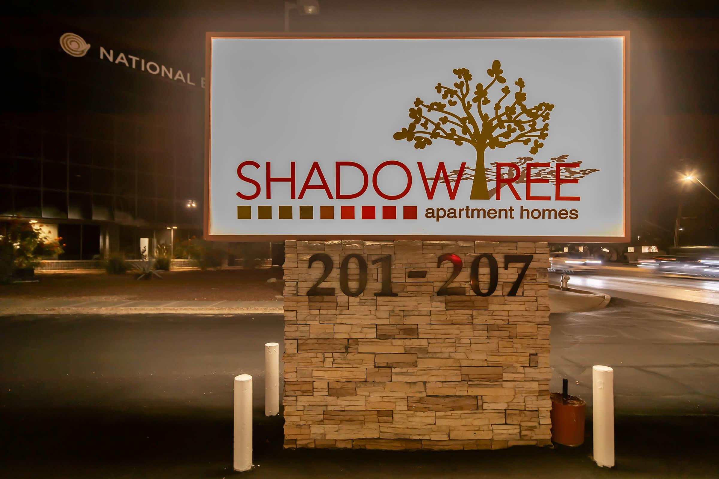 Sign for "Shadow Tree Apartment Homes" displaying the address 201-207, featuring a tree graphic. The background includes a building and street scenes at night, with illuminated signage and vehicle lights.