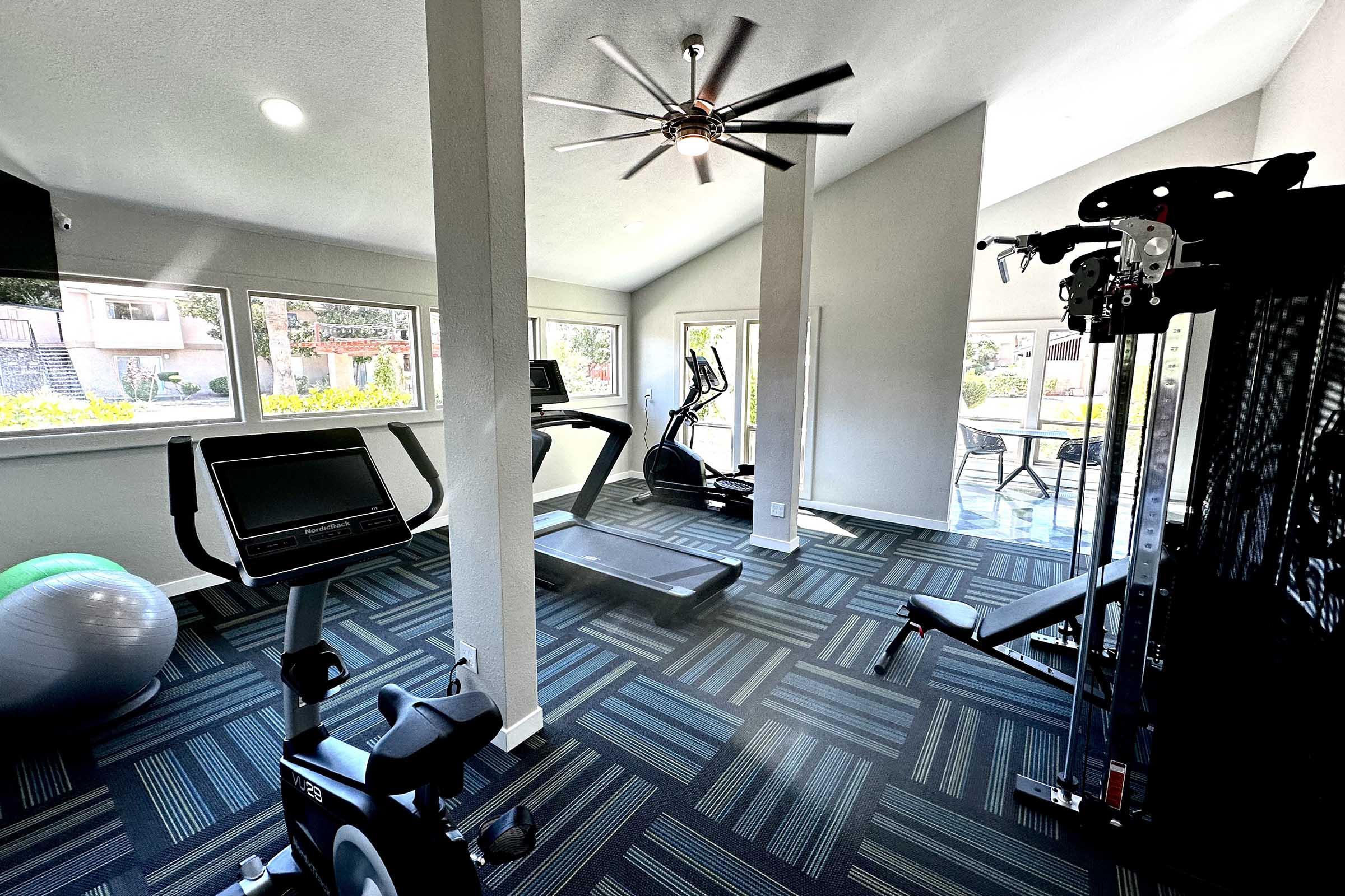 A bright and spacious gym with large windows, featuring a treadmill, stationary bike, and elliptical machine. There's a weight machine and an exercise ball, all set on a modern, patterned carpet. The ceiling has a ceiling fan, enhancing the open atmosphere.