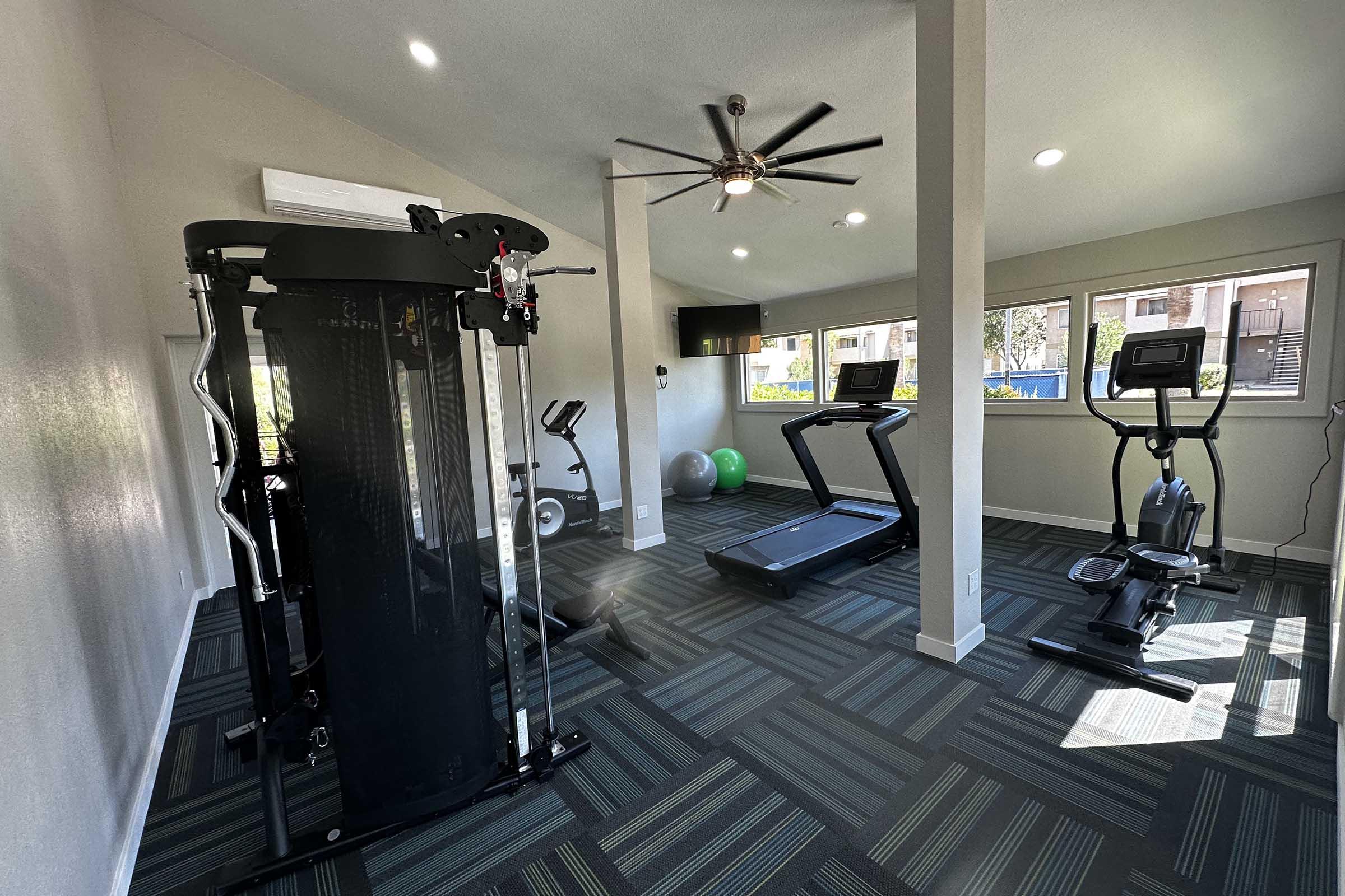 A modern gym interior featuring a multi-gym strength training machine, a treadmill, an elliptical trainer, and an exercise bike. The space is well-lit with large windows, a ceiling fan, and a green exercise ball. The flooring has a geometric pattern, enhancing the contemporary design.