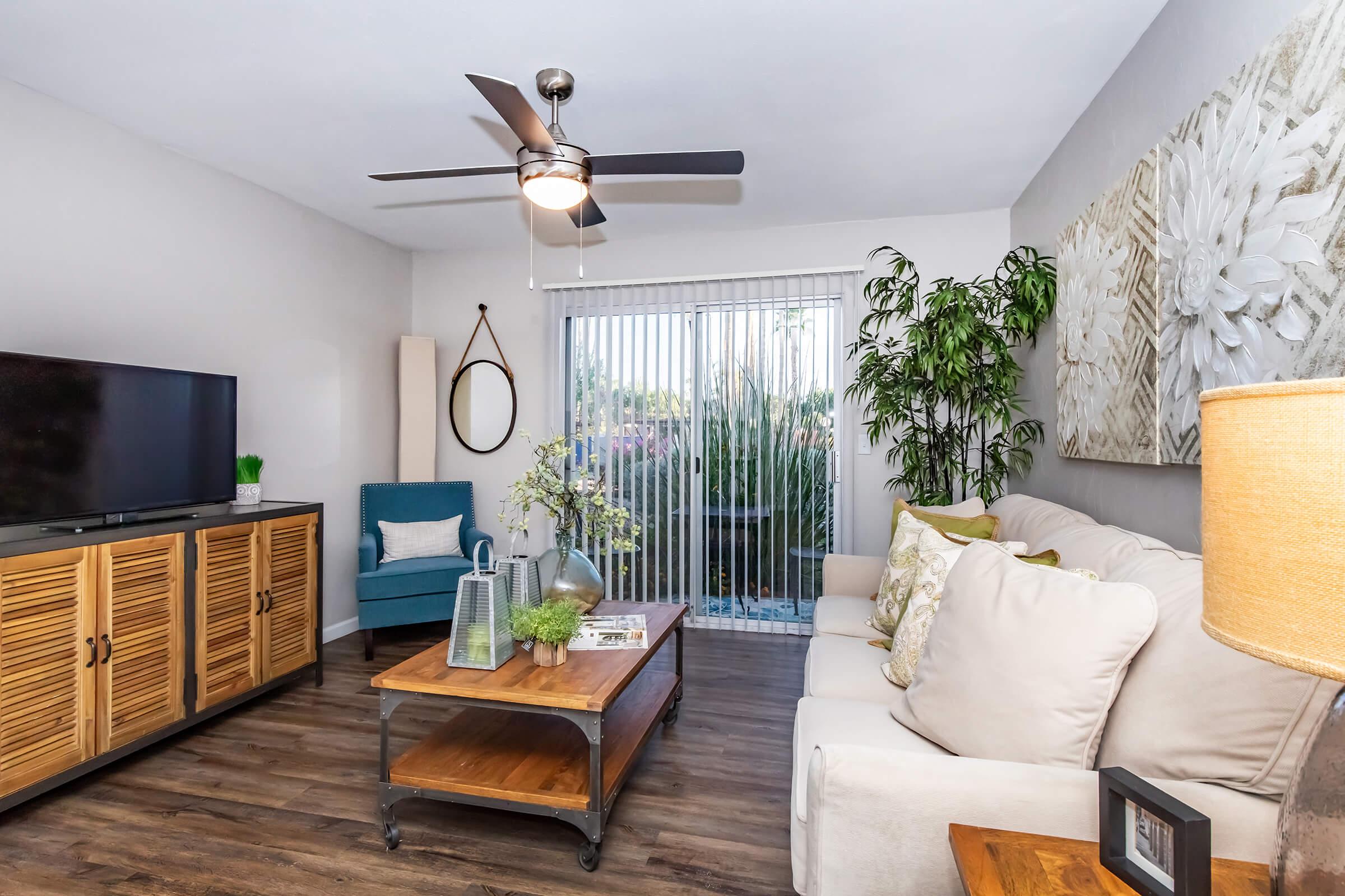 A modern living room featuring a light-colored sofa with decorative pillows, a wooden coffee table, a TV on a cabinet, and an accent chair. Large sliding glass doors allow natural light, revealing greenery outside. Decor includes a plant, wall art, and a floor lamp, creating a cozy and inviting atmosphere.