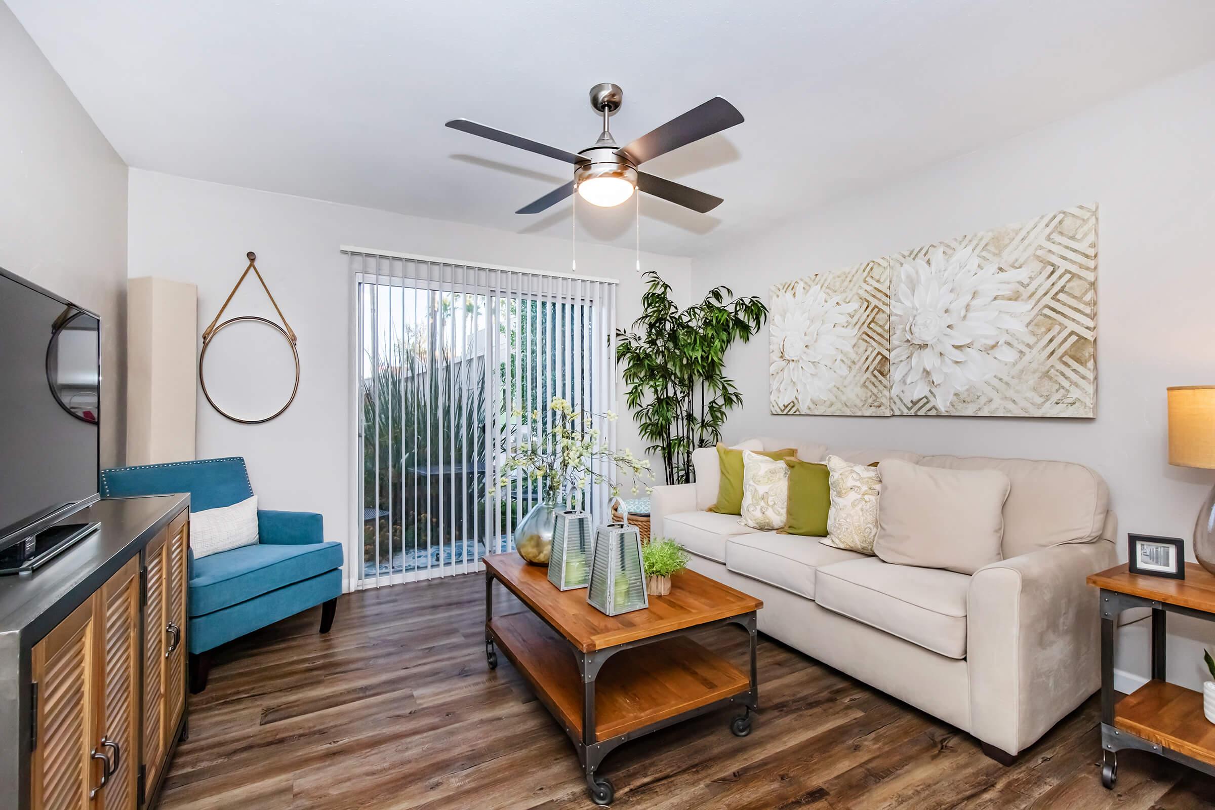A cozy living room featuring a beige sofa with decorative pillows, a wooden coffee table with green vases, and a blue accent chair. There is a ceiling fan, a television on a cabinet, and large windows with vertical blinds allowing natural light. Decorative plants enhance the cheerful ambiance.