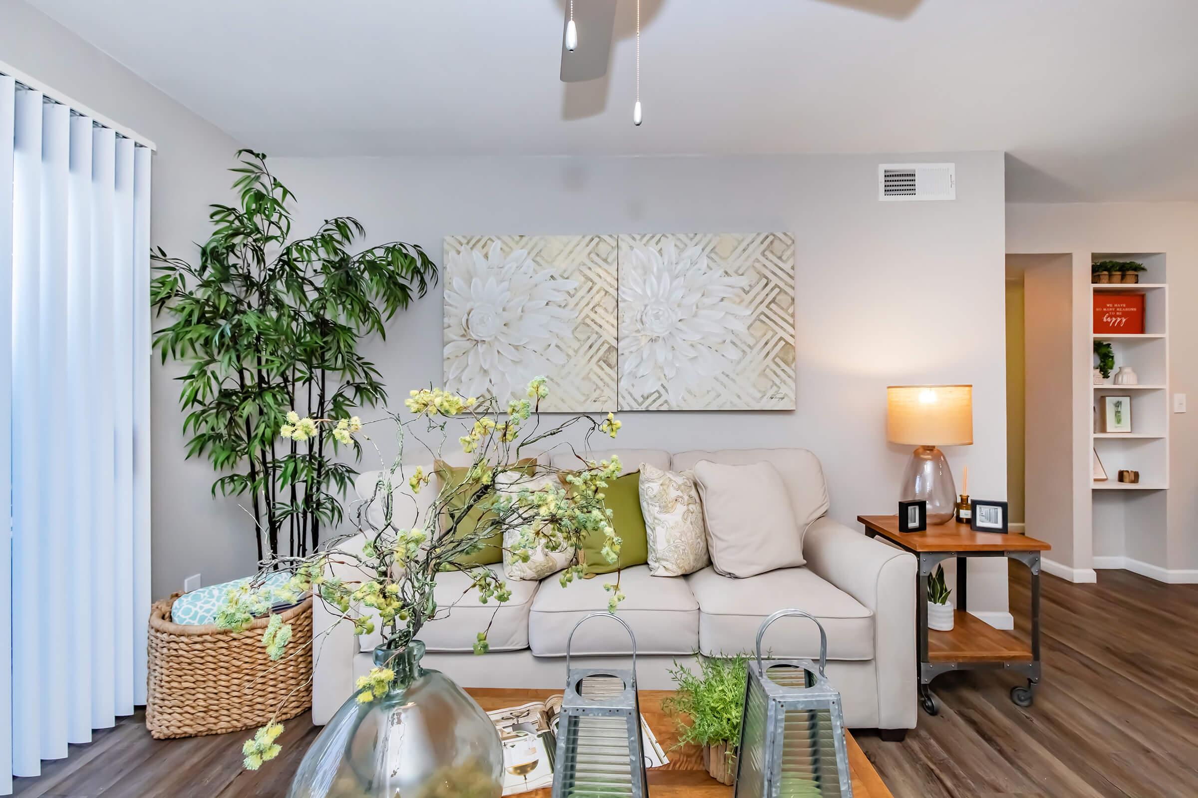 A cozy living room featuring a light-colored sofa with green and patterned cushions, a wooden coffee table with decorative items, and a floor lamp. A large artwork hangs on the wall, and there are potted plants and a woven basket nearby. Natural light filters through sheer vertical blinds.