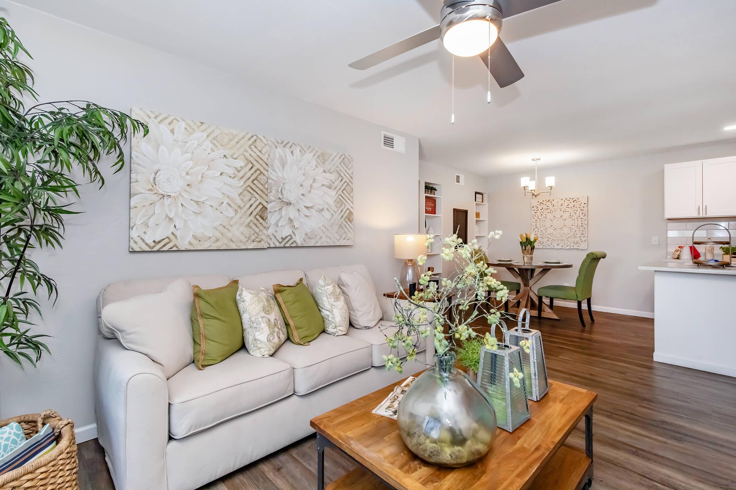Cozy living room featuring a light-colored sofa with green cushions, a wooden coffee table with decorative plants, and artwork of flowers on the wall. A kitchen area is visible in the background with a dining table and chairs. Natural light enhances the warm and inviting atmosphere.