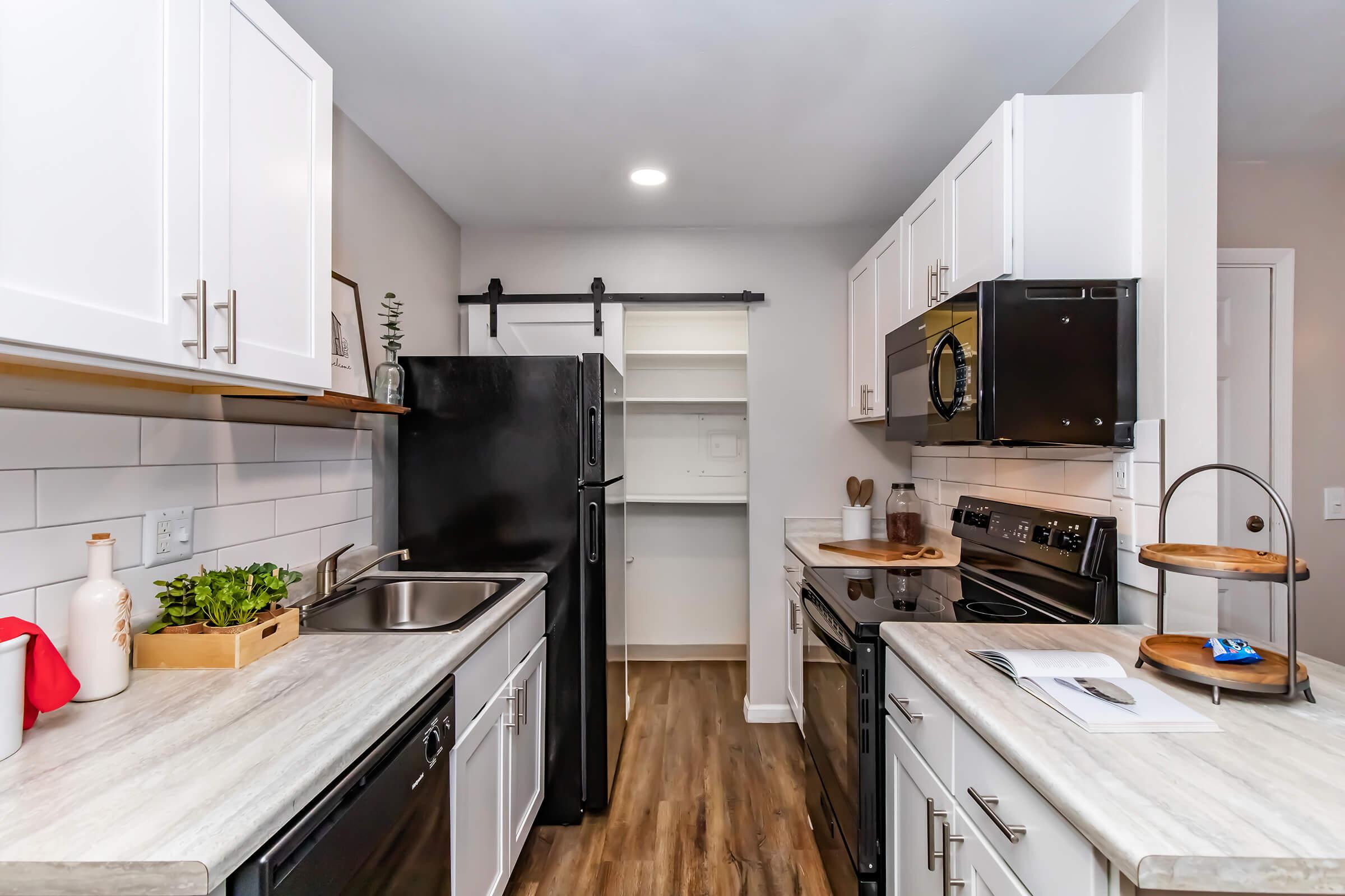 Modern kitchen featuring white cabinets, stainless steel appliances, a dark refrigerator, and a spacious countertop. The design includes a farm-style sink, wooden accents, and an open pantry area. The floor is a warm-toned wood laminate, creating a cozy atmosphere. Bright overhead lighting enhances the airy feel.