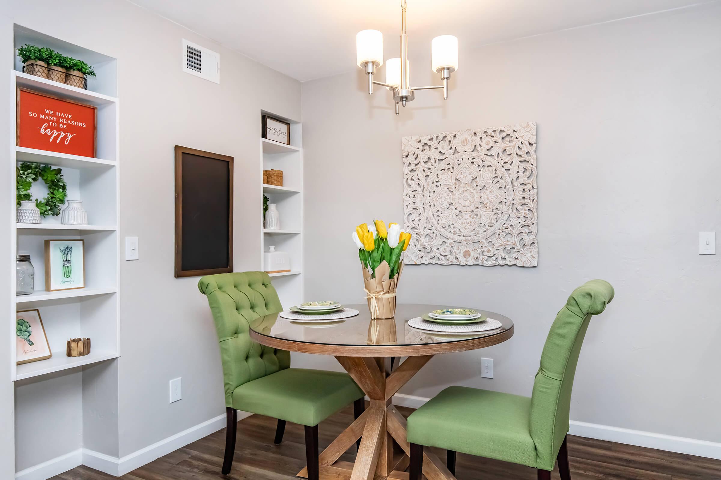 A cozy dining area featuring a round wooden table with a vase of yellow tulips at the center, surrounded by two green upholstered chairs. The walls are decorated with a large white wall art piece and shelves containing decorative items and plants, creating a welcoming ambiance.