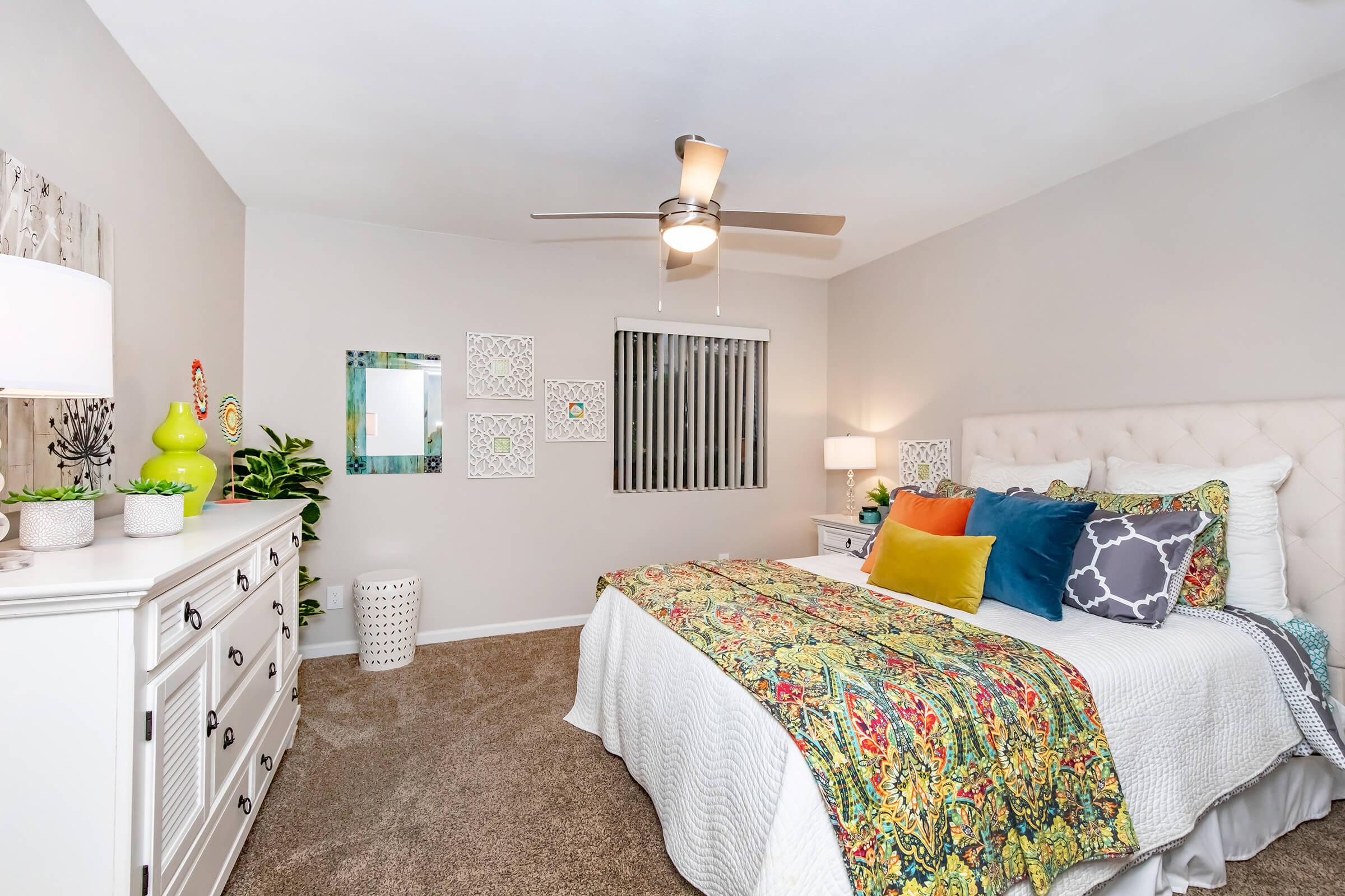 A cozy bedroom featuring a neatly made king-sized bed with a vibrant patterned quilt and colorful pillows. There is a white dresser, a small table, and decorative plants. A ceiling fan is overhead, and the walls are adorned with framed art. Natural light filters through a window with blinds.