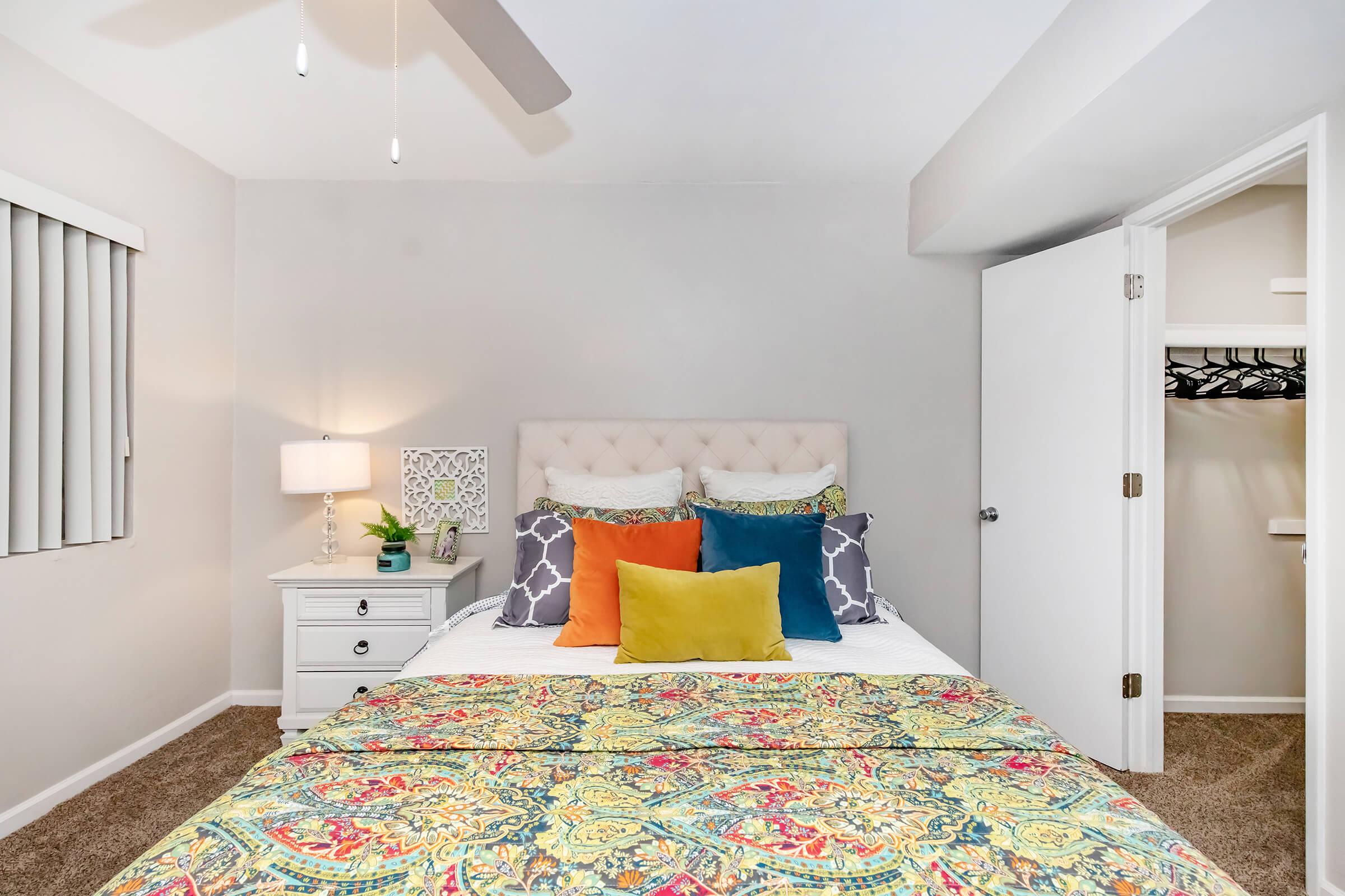 Cozy bedroom featuring a queen-sized bed with colorful pillows and a patterned bedspread. A nightstand with a lamp is beside the bed, and there's a closet with wire shelving visible through an open door. The walls are painted in a soft gray, enhancing the room's inviting atmosphere.