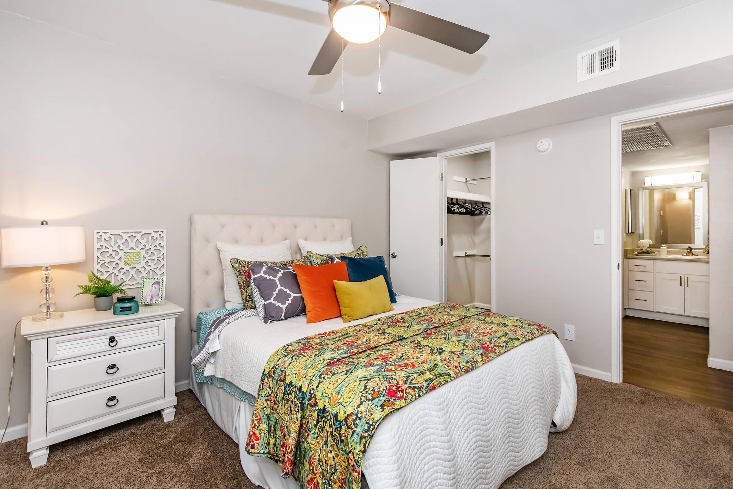 A cozy bedroom featuring a queen-sized bed with colorful decorative pillows and a patterned bedspread. A nightstand with a lamp sits beside the bed, and there's a wardrobe with a clothes rack in the closet. The walls are painted light gray, and there's a ceiling fan for ventilation.