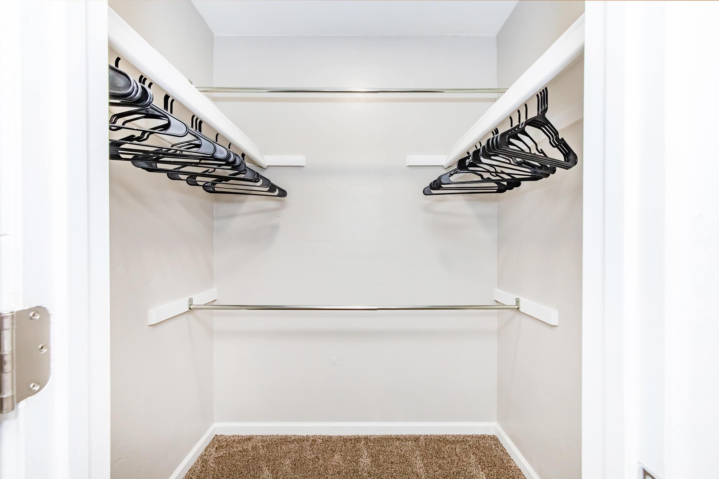 A spacious, empty closet with two rows of silver clothing rods and several black hangers lined up. The walls are light-colored, and the floor is covered with a soft, tan carpet. The closet has a clean and organized appearance, providing ample storage space.