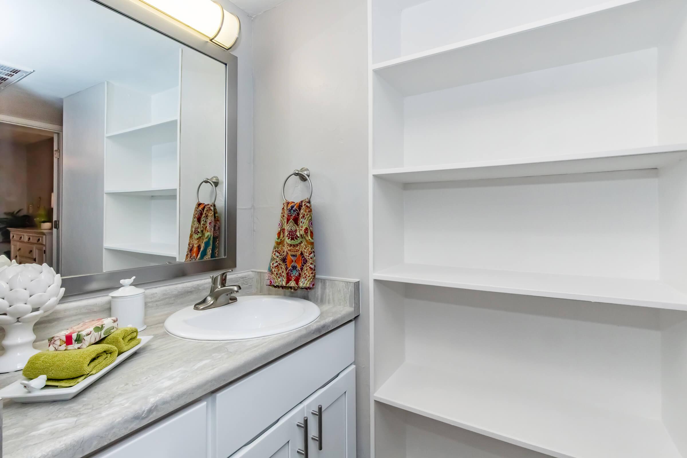 A bright bathroom with a white countertop, a round sink, and a large mirror above it. On the counter, there are decorative items: a white ceramic dish, a small container, and a green towel. Two shelves are mounted on the wall, one fully empty and the other displaying colorful towels. Natural light enters from a nearby window.