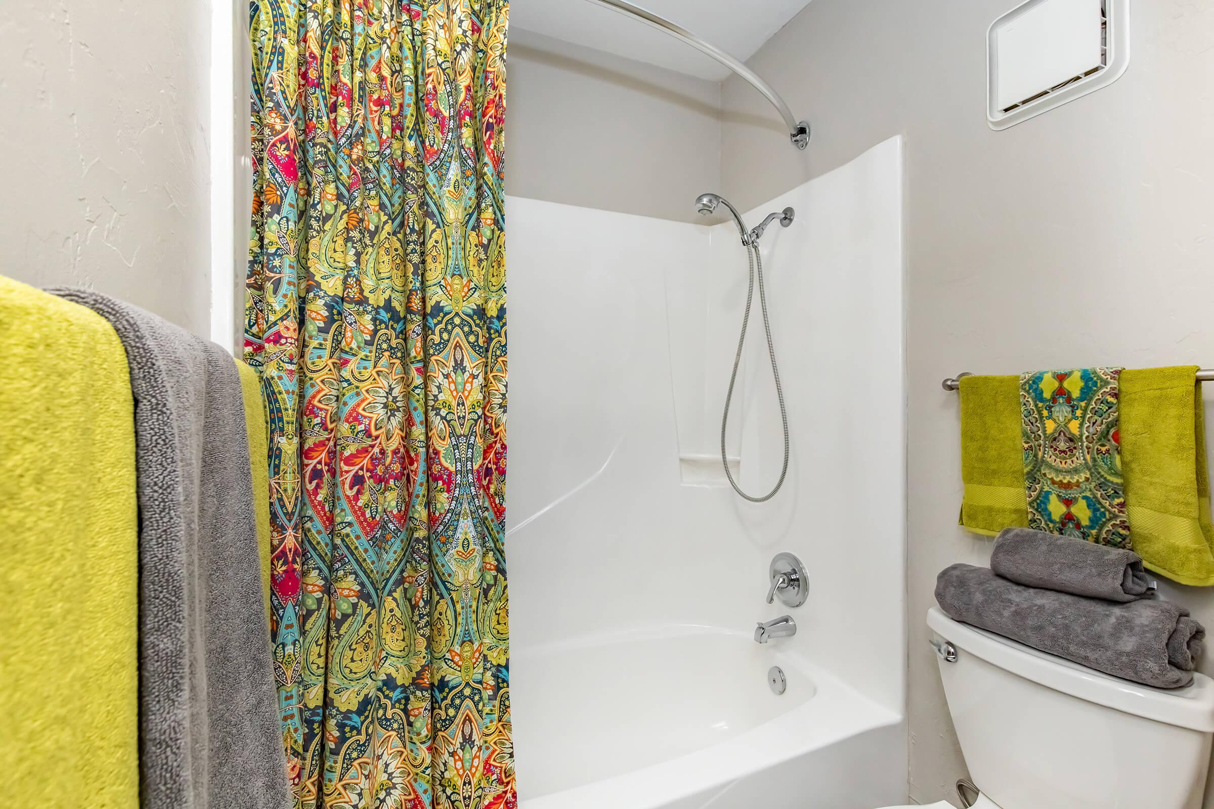 A brightly colored shower curtain with a floral pattern hangs in a bathroom. The bathtub is white and includes a detachable showerhead. Towels in gray and green are neatly placed nearby. The walls are a muted color, creating a clean and inviting atmosphere.