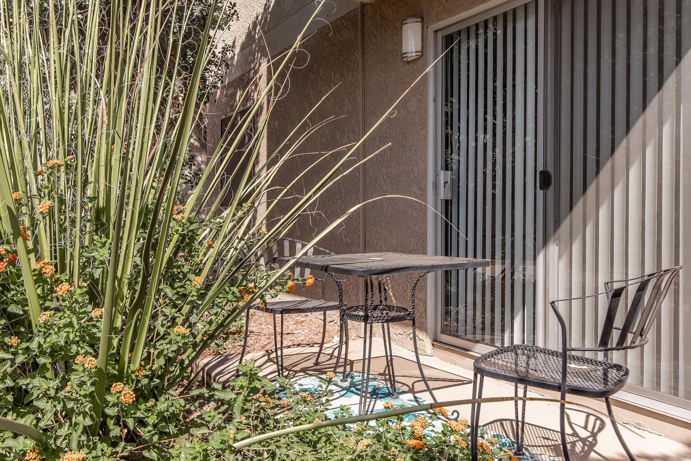 A small outdoor patio featuring a black metal table and two chairs, surrounded by lush green plants and colorful flowers. A sliding glass door with vertical blinds is in the background, capturing a sunny and inviting atmosphere.