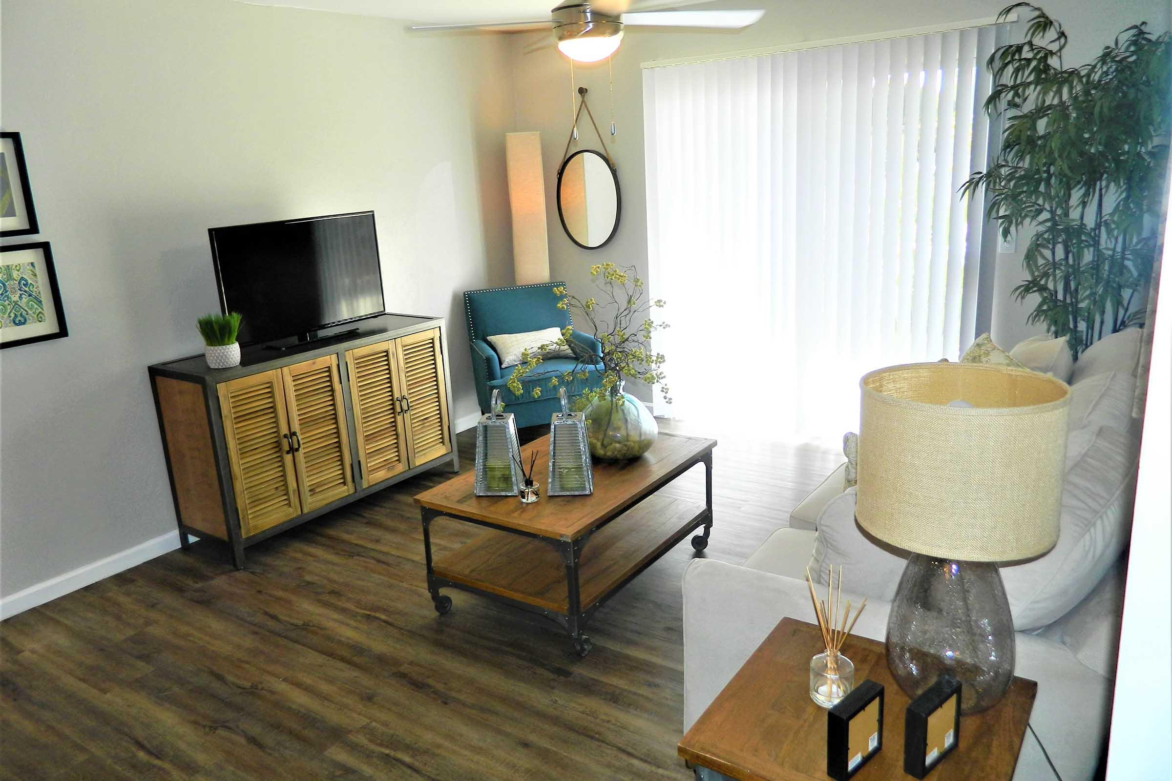 A cozy living room featuring a TV on a wooden cabinet, a comfortable blue chair, a coffee table with decorative items, and a plant. Soft lighting from a lamp and large windows with blinds create a warm atmosphere. There are also green plants for added decor.