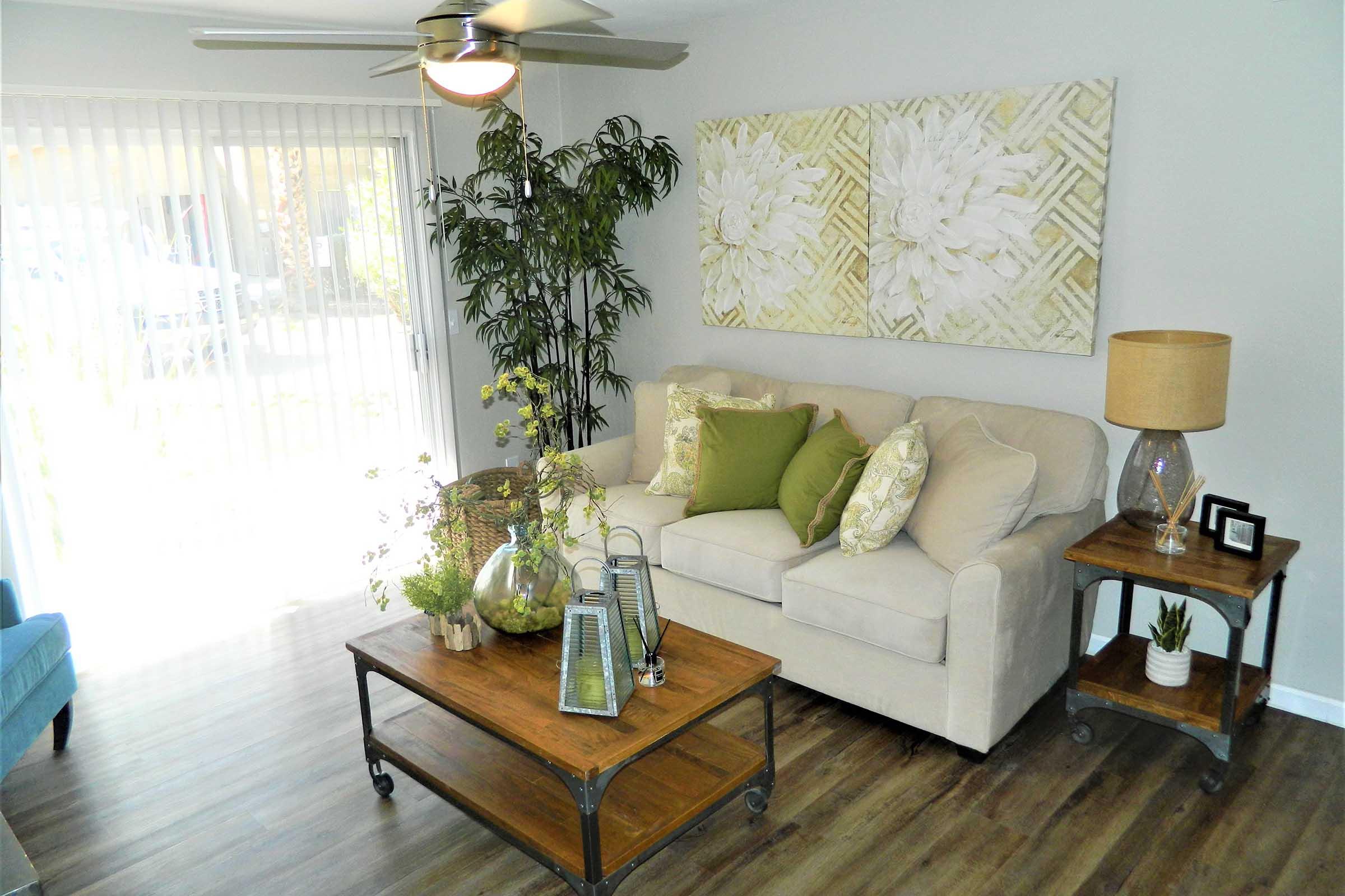 A cozy living room featuring a light-colored sofa with decorative pillows, a wooden coffee table with plants and decorative items, and a floor lamp. Large windows with vertical blinds allow natural light to fill the space, complemented by a leafy plant and a wall art piece depicting floral designs.