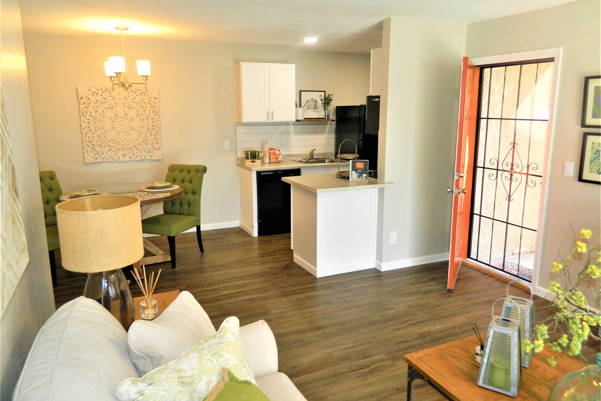 A modern living space featuring a white sofa with decorative pillows, a wooden coffee table, and a lamp. In the background, there's a small dining area with a round table and green chairs, a kitchenette with white cabinets, and a bright orange front door. Natural light fills the room, creating a welcoming atmosphere.