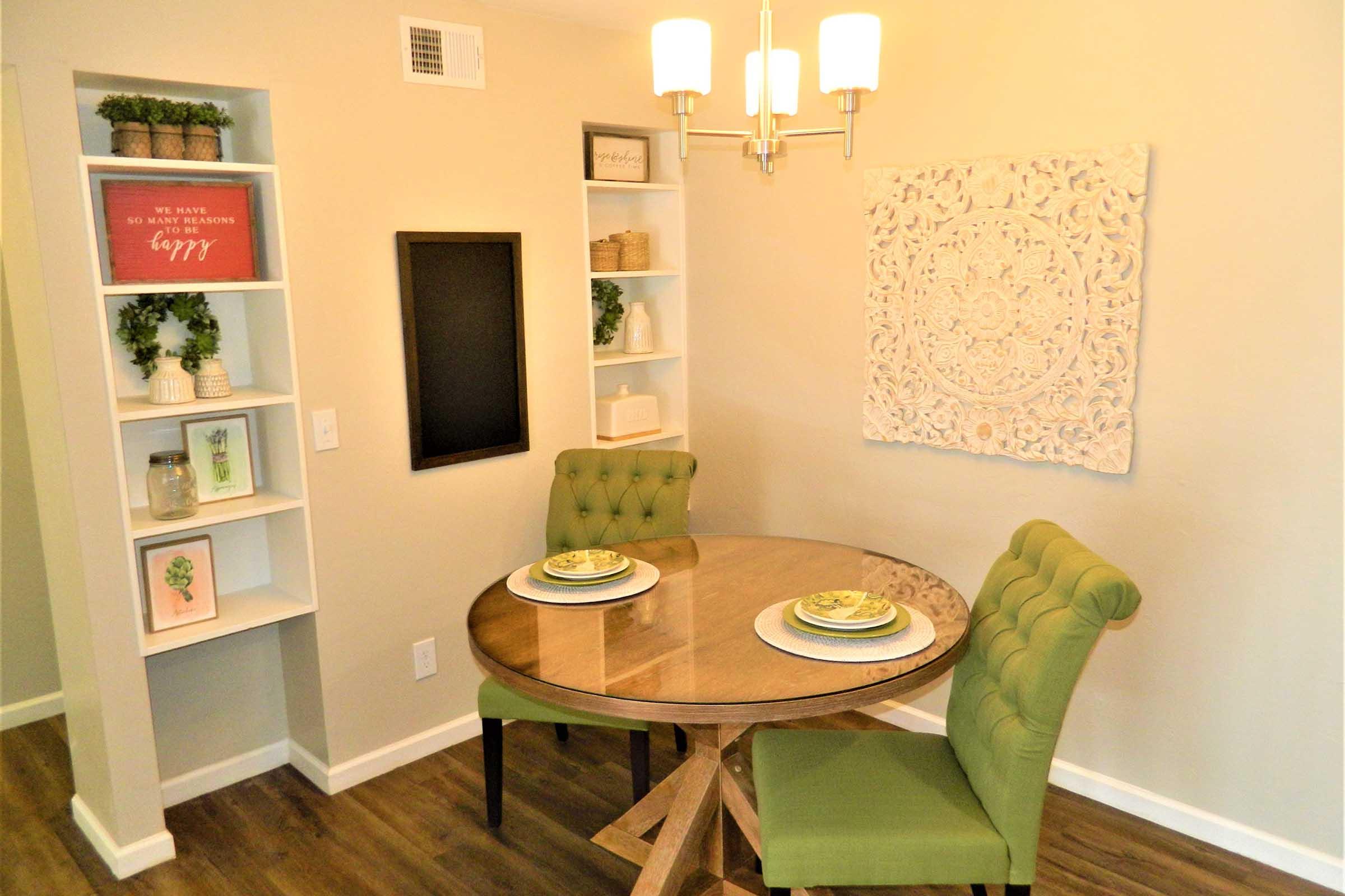 A cozy dining area featuring a round wooden table with two green chairs. The table is set with plates and decorative items. A light fixture hangs above, and the walls are adorned with a decorative wall hanging and a black chalkboard. Shelves with plants and decorations are in the background.