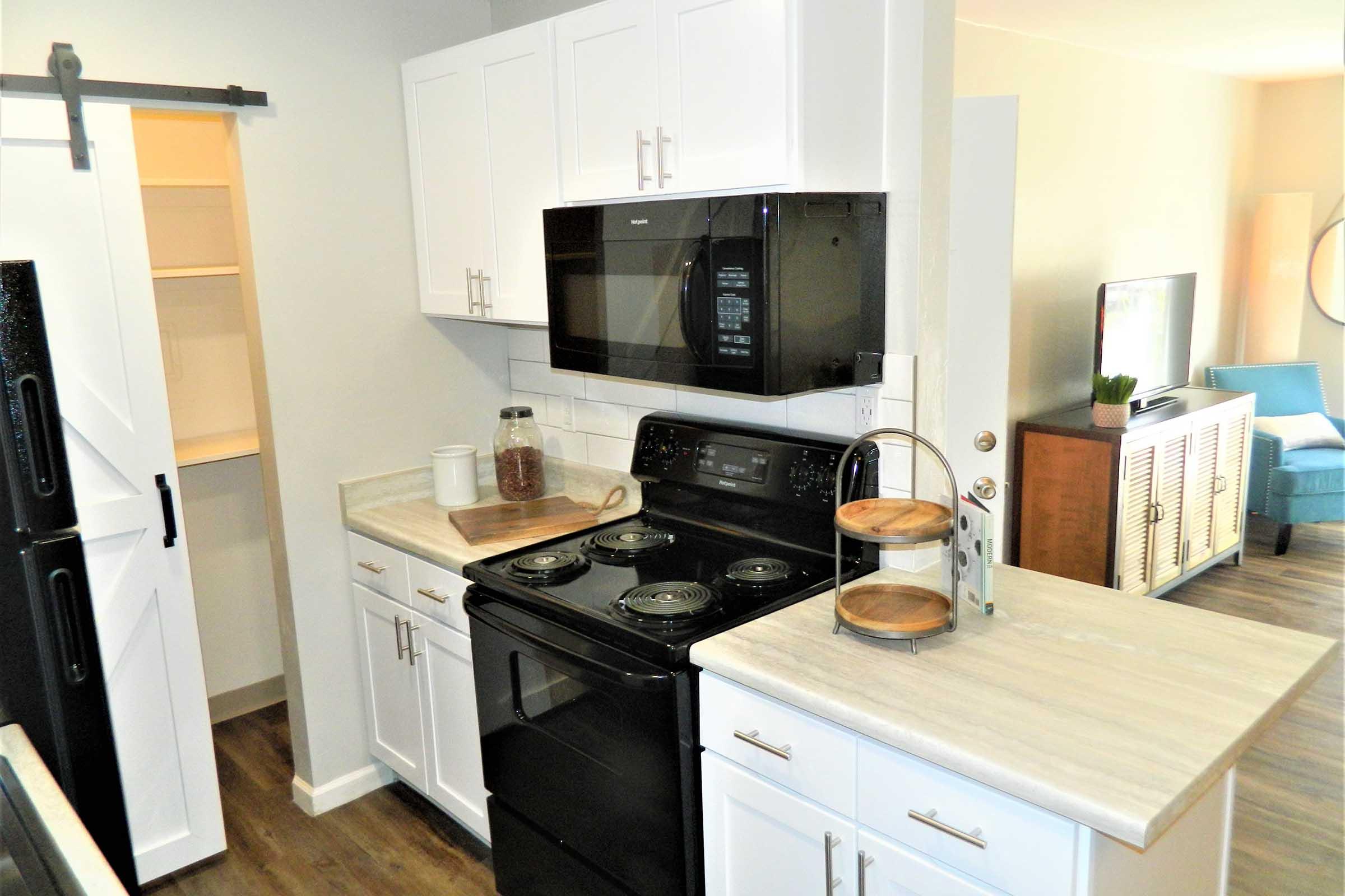 A modern kitchen featuring white cabinets, a black stove, and microwave. There's a wooden countertop with a tray of snacks and a corkscrew. The refrigerator is black, and shelves are visible in the background. A light-colored living area with a blue chair and TV can be seen beyond the kitchen.