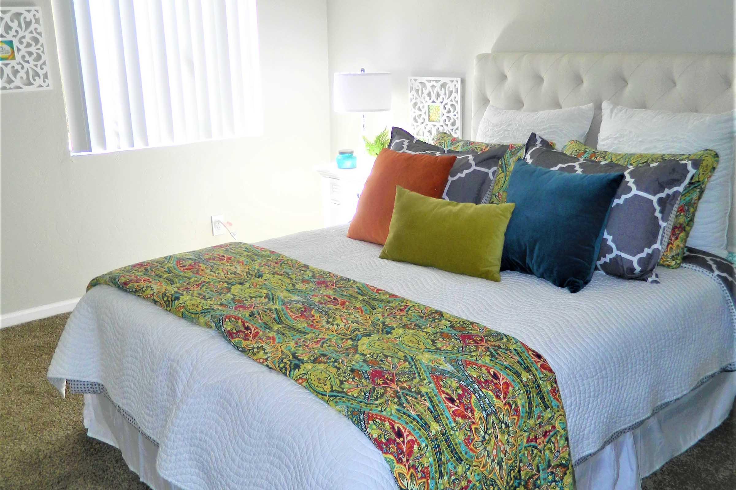 A cozy bedroom featuring a neatly made bed with a white quilt and a colorful decorative throw. The bed is adorned with various pillows in different shades, including blue, orange, and green. Soft natural light filters through a window with vertical blinds, creating a warm atmosphere in the room.