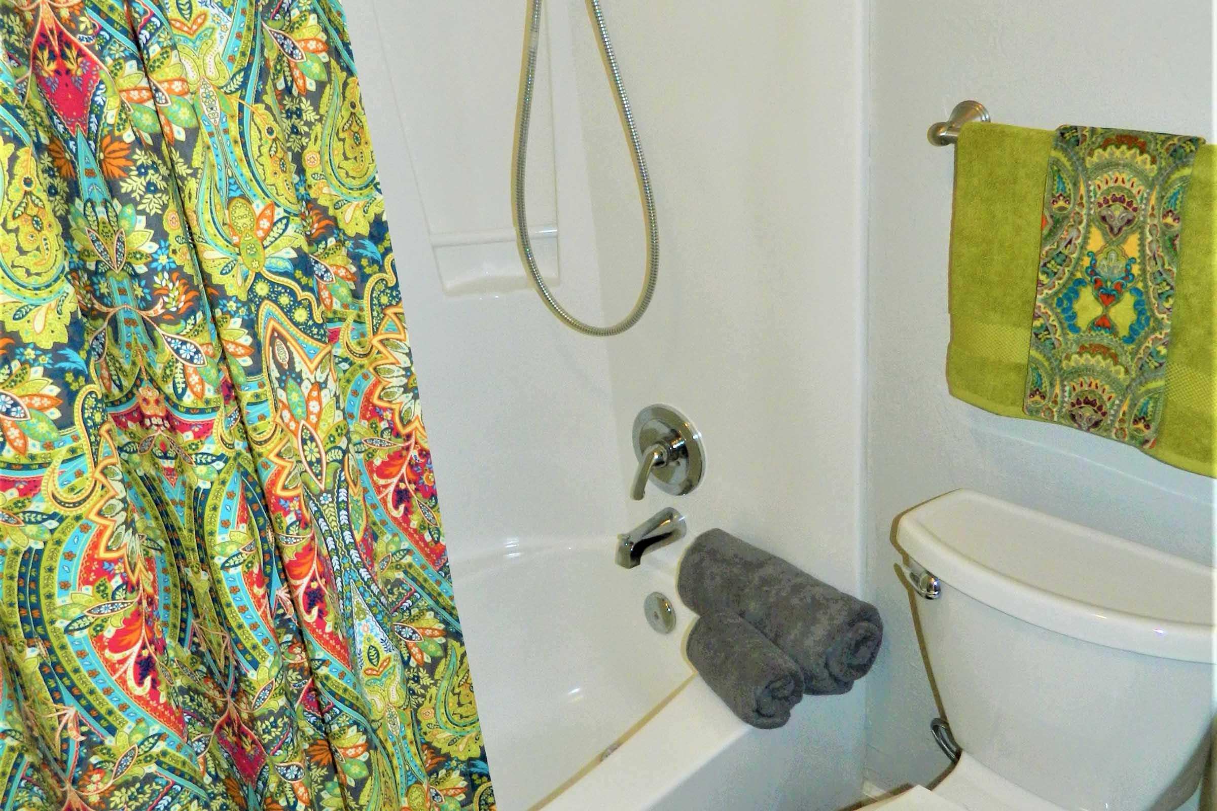A colorful shower curtain with an intricate pattern hangs in a bathroom. There is a showerhead, a bathtub, a toilet with neatly rolled gray towels beside it, and a green hand towel on the wall. The overall atmosphere is bright and vibrant, showcasing a decorative and functional bathroom space.