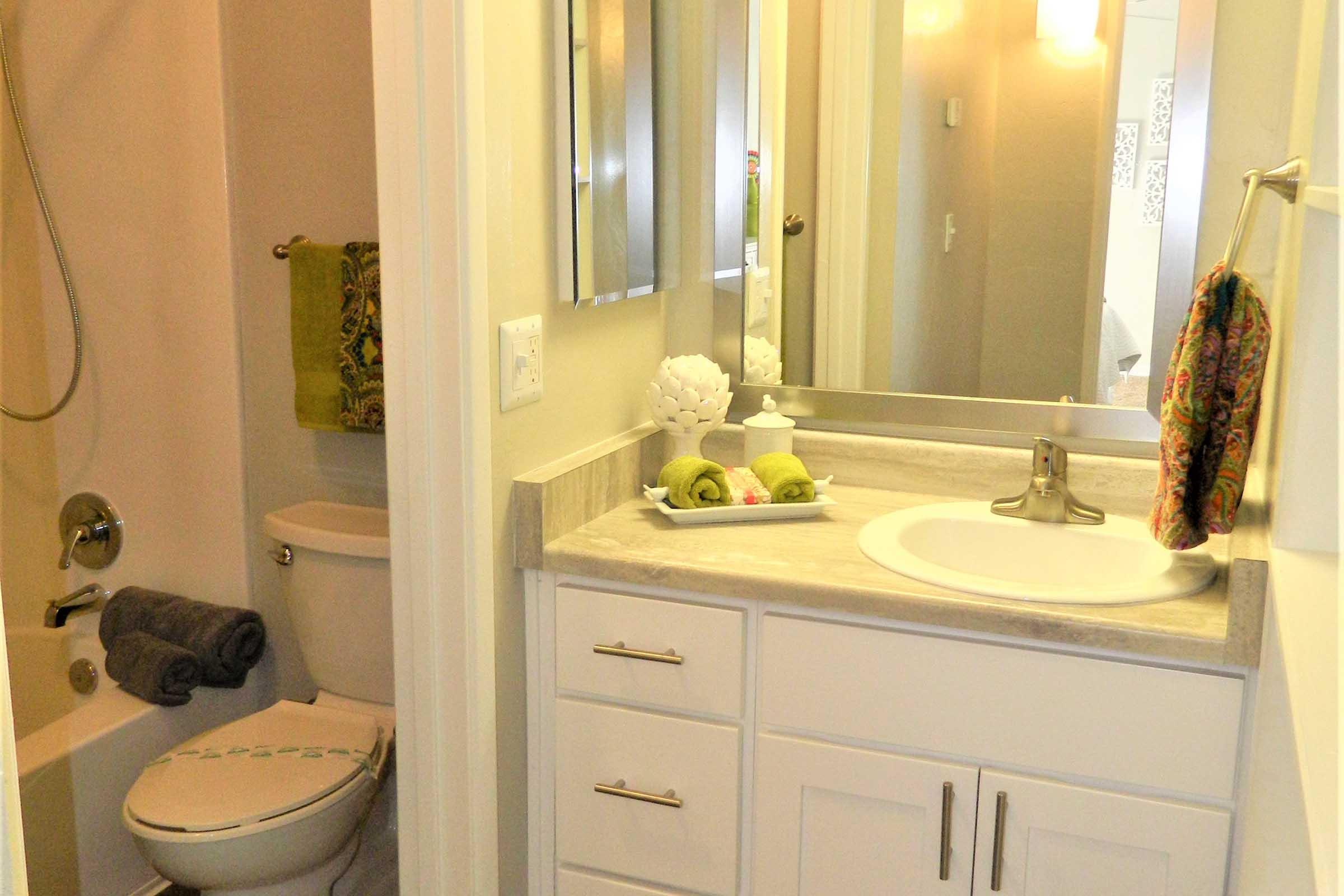 A small, modern bathroom featuring a shower with a glass door, a sink with a granite countertop, and white cabinetry. Towels are neatly arranged on the countertop, and decorative items, such as a flower and green towels, add a fresh touch. The walls are painted in neutral tones.