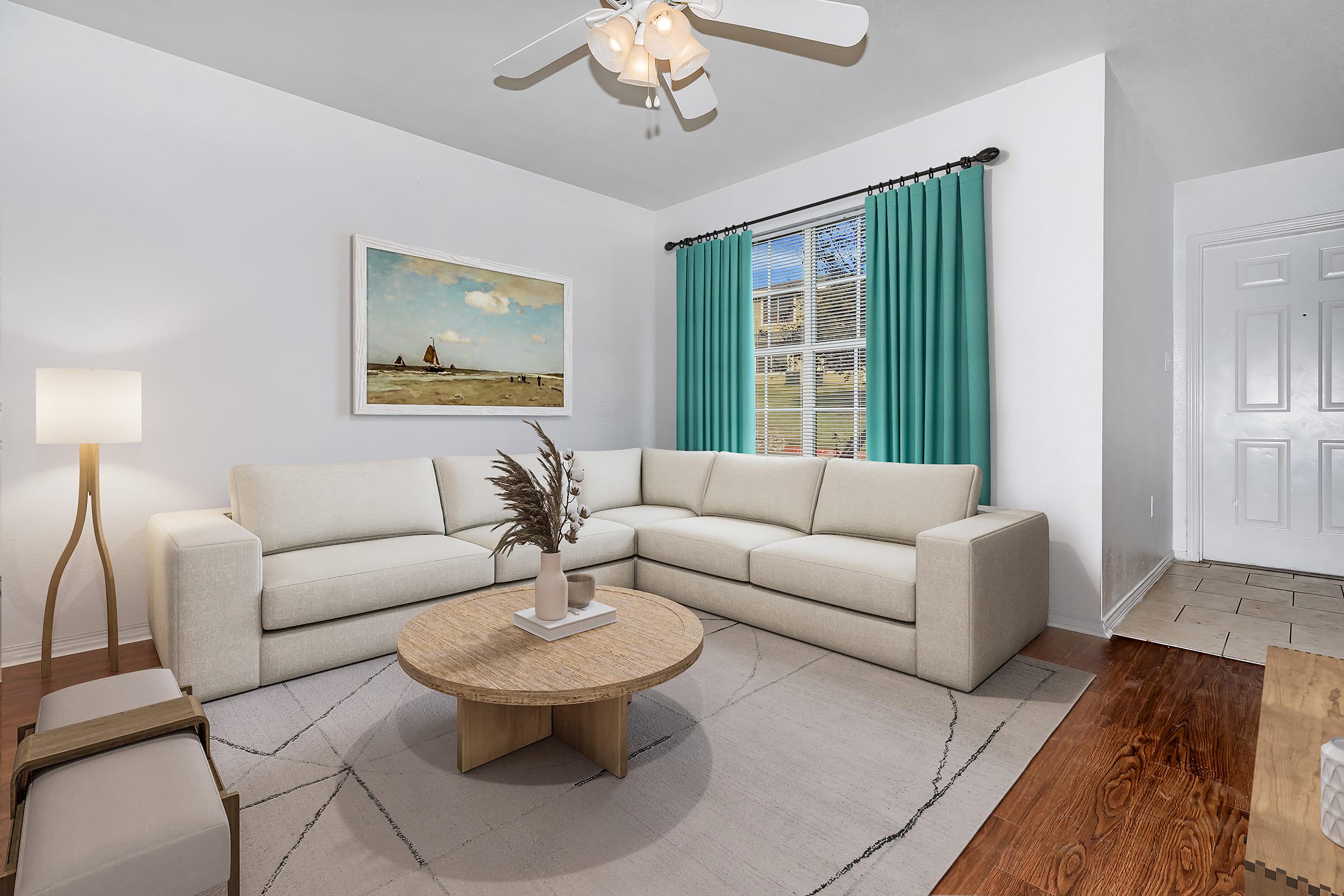A modern living room featuring a light-colored sectional sofa, a round wooden coffee table, and a decorative plant. There is an artwork with sailboats on the wall and large windows with turquoise curtains, allowing natural light to fill the space. The flooring is a mix of wood and tile.