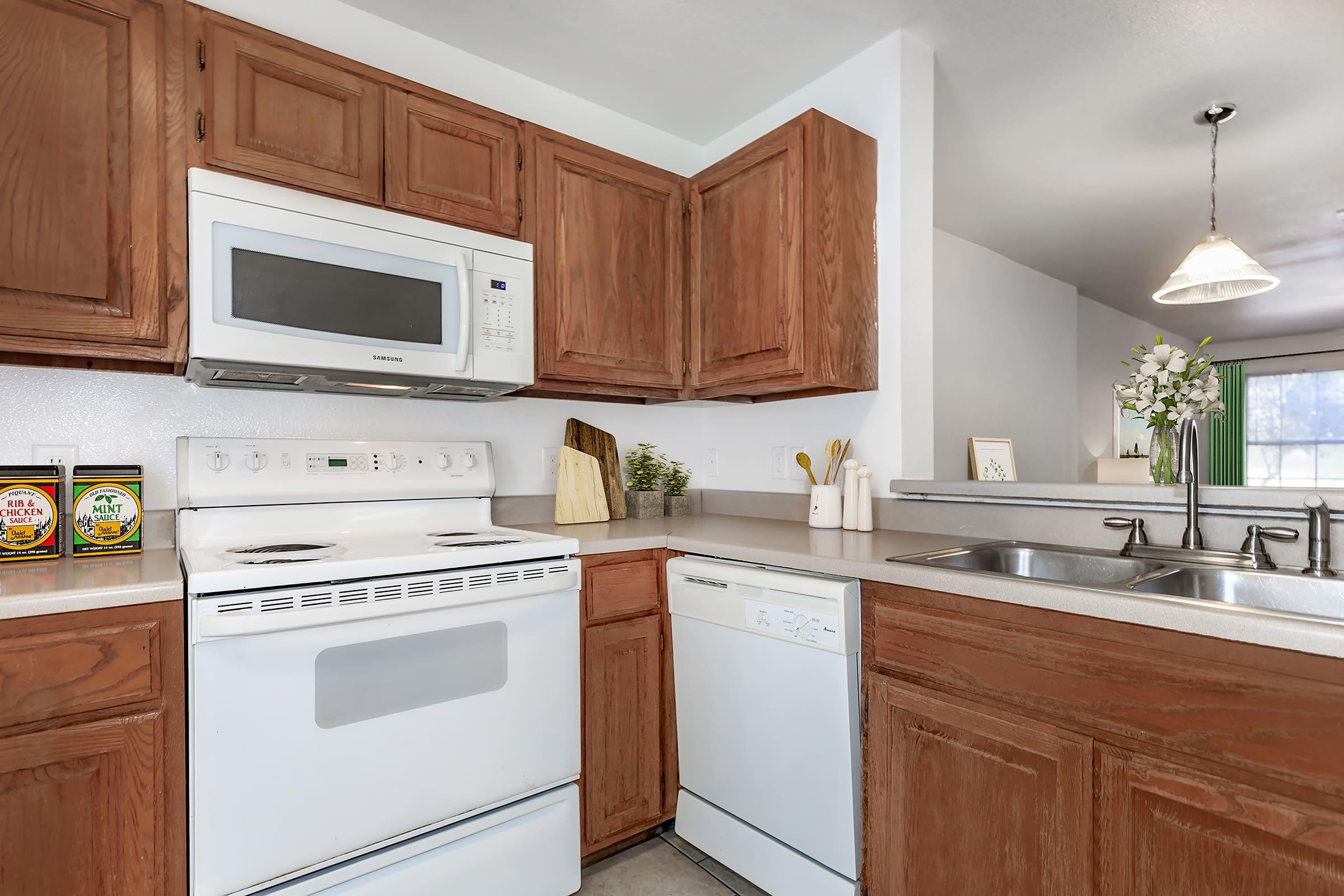 A modern kitchen featuring wooden cabinetry, a white microwave above a stove, and a dishwasher. The countertop is clean, with a small plant and kitchen accessories. Bright lighting from a nearby window enhances the inviting atmosphere.