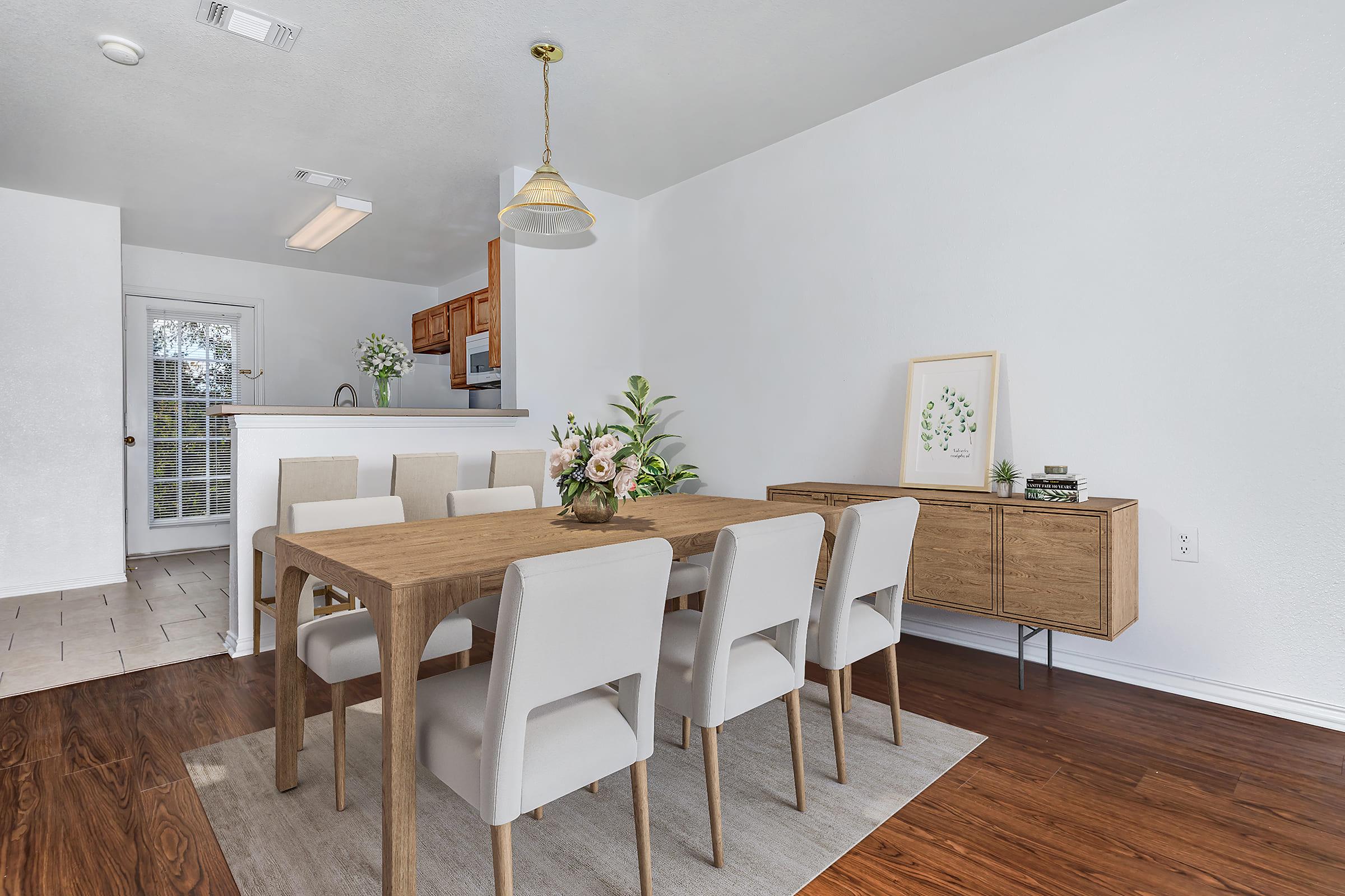 A spacious dining area featuring a large wooden table surrounded by light-colored upholstered chairs. A modern pendant light hangs above the table. A sideboard with decorative items is positioned against the wall, and a potted plant adds a touch of greenery. The space has a bright, airy feel with light-colored walls and hardwood flooring.