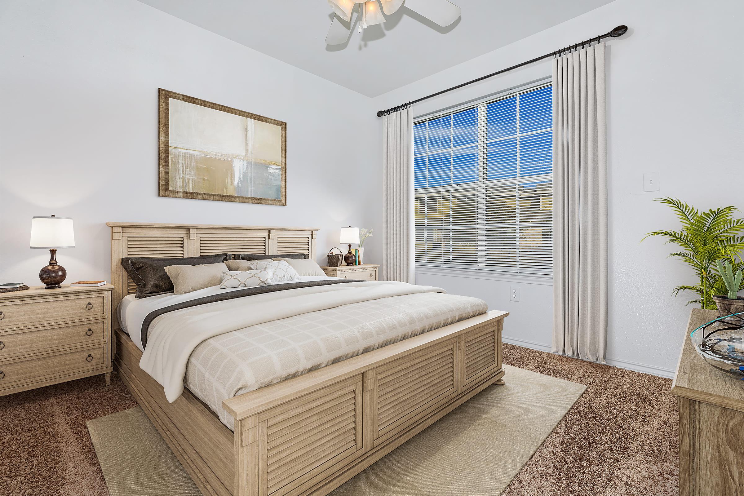 A cozy bedroom featuring a wooden bed with soft bedding, two bedside lamps, a wooden dresser, and a large window with sheer curtains. The walls are painted white, and there's a piece of abstract art above the bed. A potted plant adds a touch of greenery to the space. Sunlight streams in through the window.