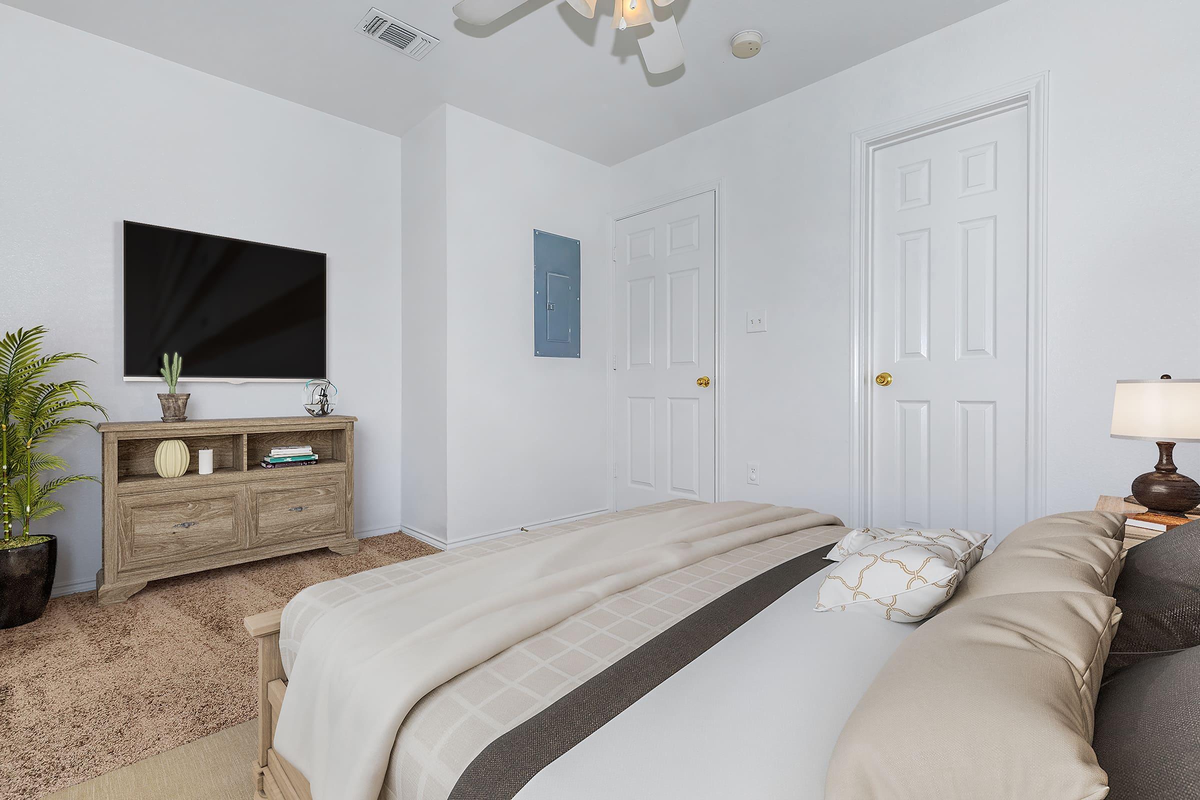 A cozy bedroom featuring a neatly made bed with decorative pillows, a wooden nightstand with a lamp, a TV mounted on the wall, and a small plant on a dresser. The walls are painted white, and there are two closed doors in the room, contributing to a clean and bright aesthetic.