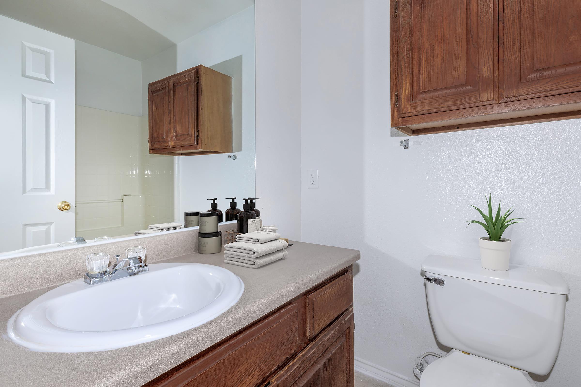 A modern bathroom featuring a clean white sink with a round basin, a wooden vanity with open shelves, and neatly arranged towels. The toilet is positioned nearby, with a small potted plant on top. A large mirror reflects the space, and light-colored walls enhance the bright, airy feel of the room.