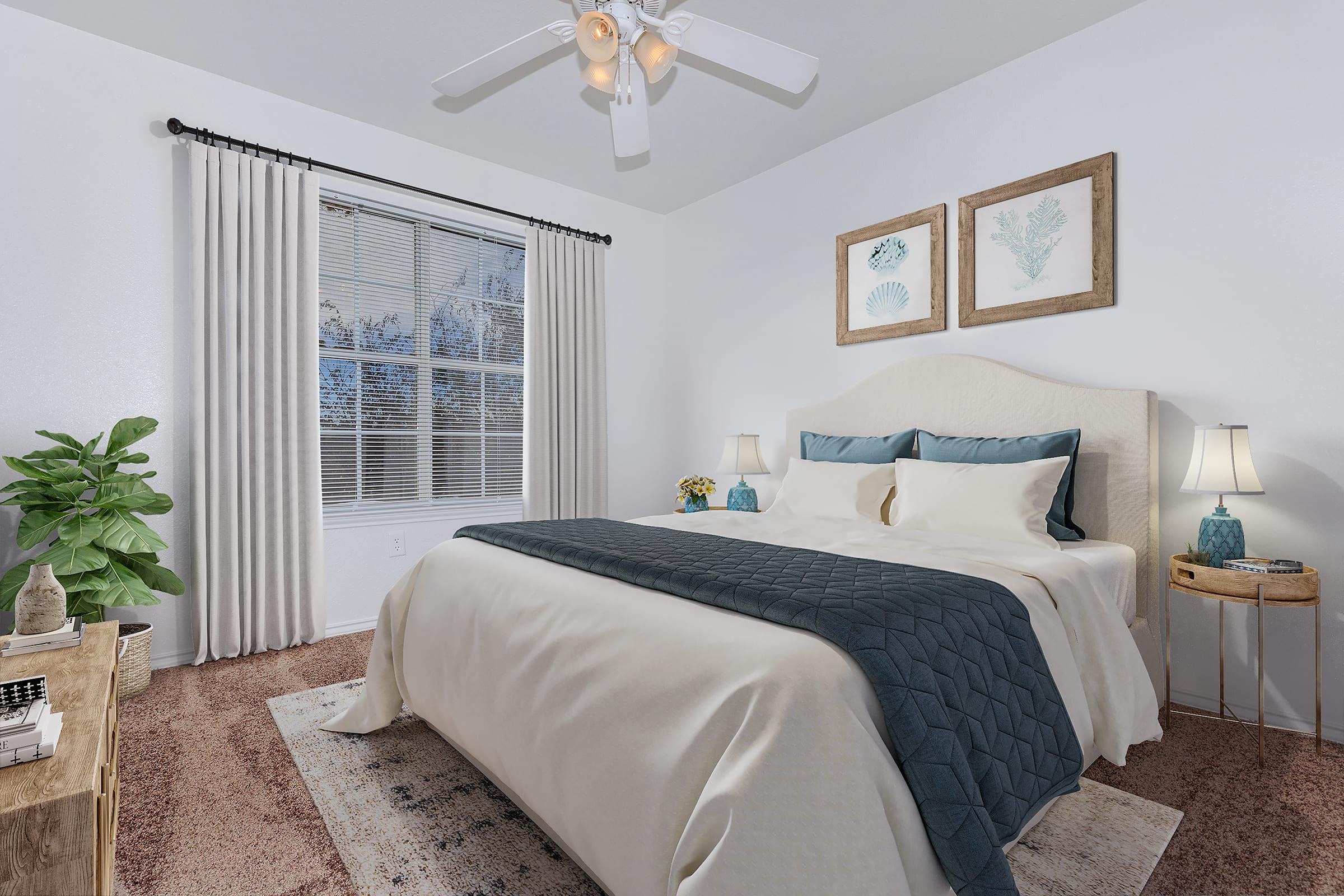 A cozy bedroom featuring a large bed with a light-colored quilt, two bedside lamps, and framed sea-themed artwork on the walls. A potted plant adds a touch of greenery. The room has a ceiling fan and soft, natural light coming through a window with sheer curtains. The carpet is a warm brown.