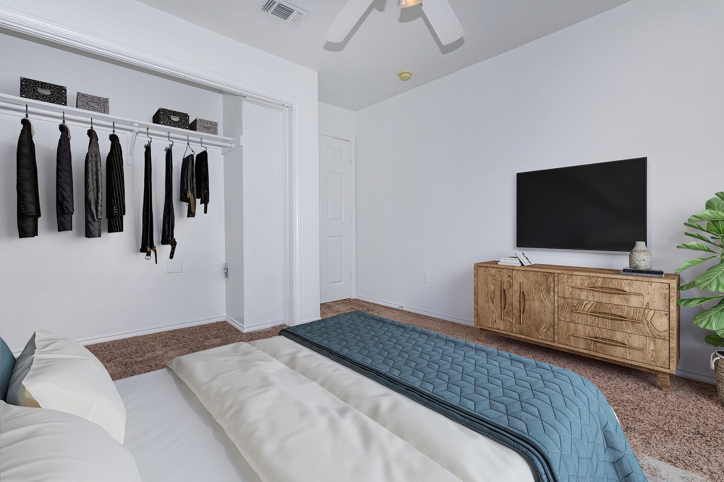A modern bedroom featuring a neatly made bed with a blue quilt. On the left, a closet with hanging clothes. A wooden dresser with a flat-screen TV sits opposite the bed, and a potted plant adds a touch of greenery to the space. The walls are painted white, creating a bright and airy feel.
