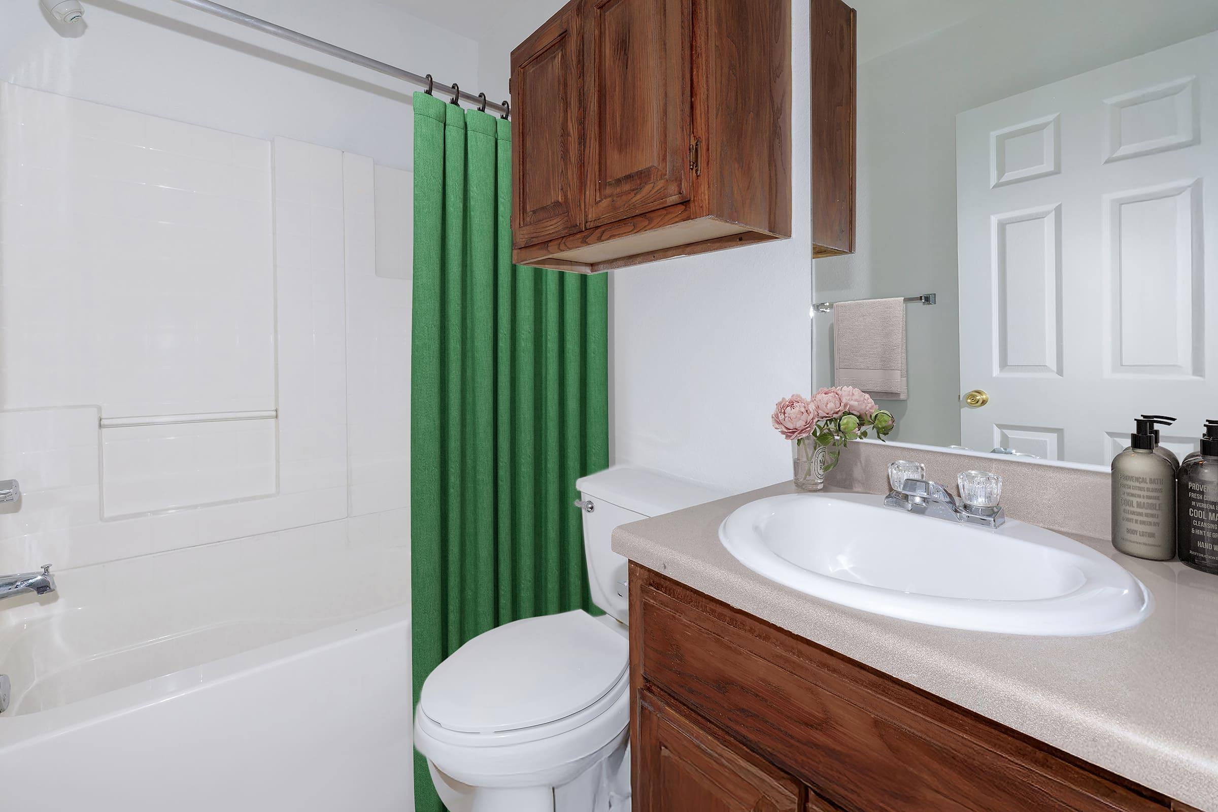 A bathroom featuring a white bathtub and shower with a green shower curtain. There is a wooden cabinet above the sink, which has a beige countertop. The sink area includes a small vase with flowers and two bottles of liquid soap. A door is visible in the background, adding depth to the space.