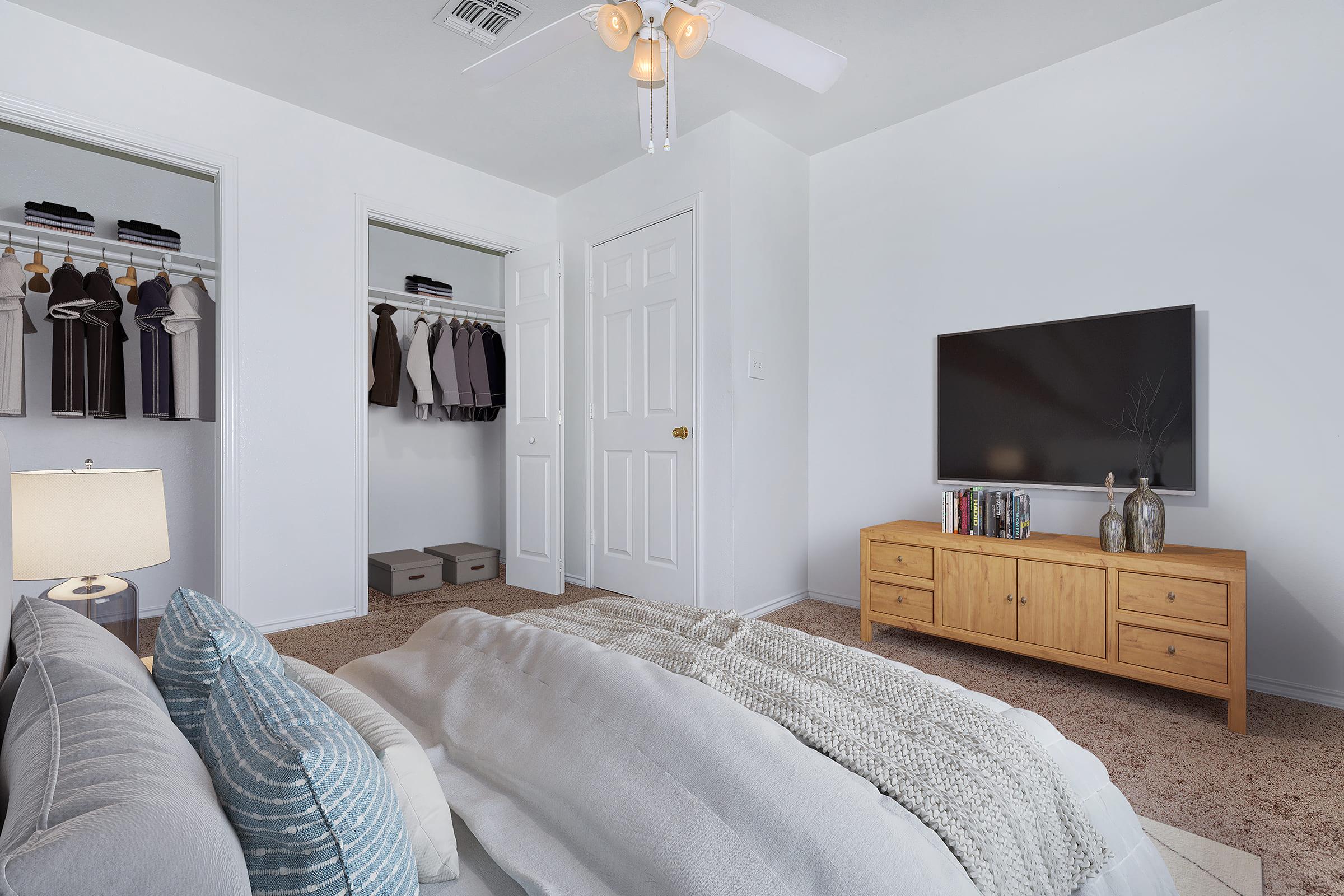 A cozy bedroom with a neatly made bed featuring textured pillows and a throw blanket. In the background, there's a closet with hanging clothes, a dresser with books and decorative items, and a wall-mounted TV. The room is well-lit with a ceiling fan and a bedside lamp, creating a warm and inviting atmosphere.