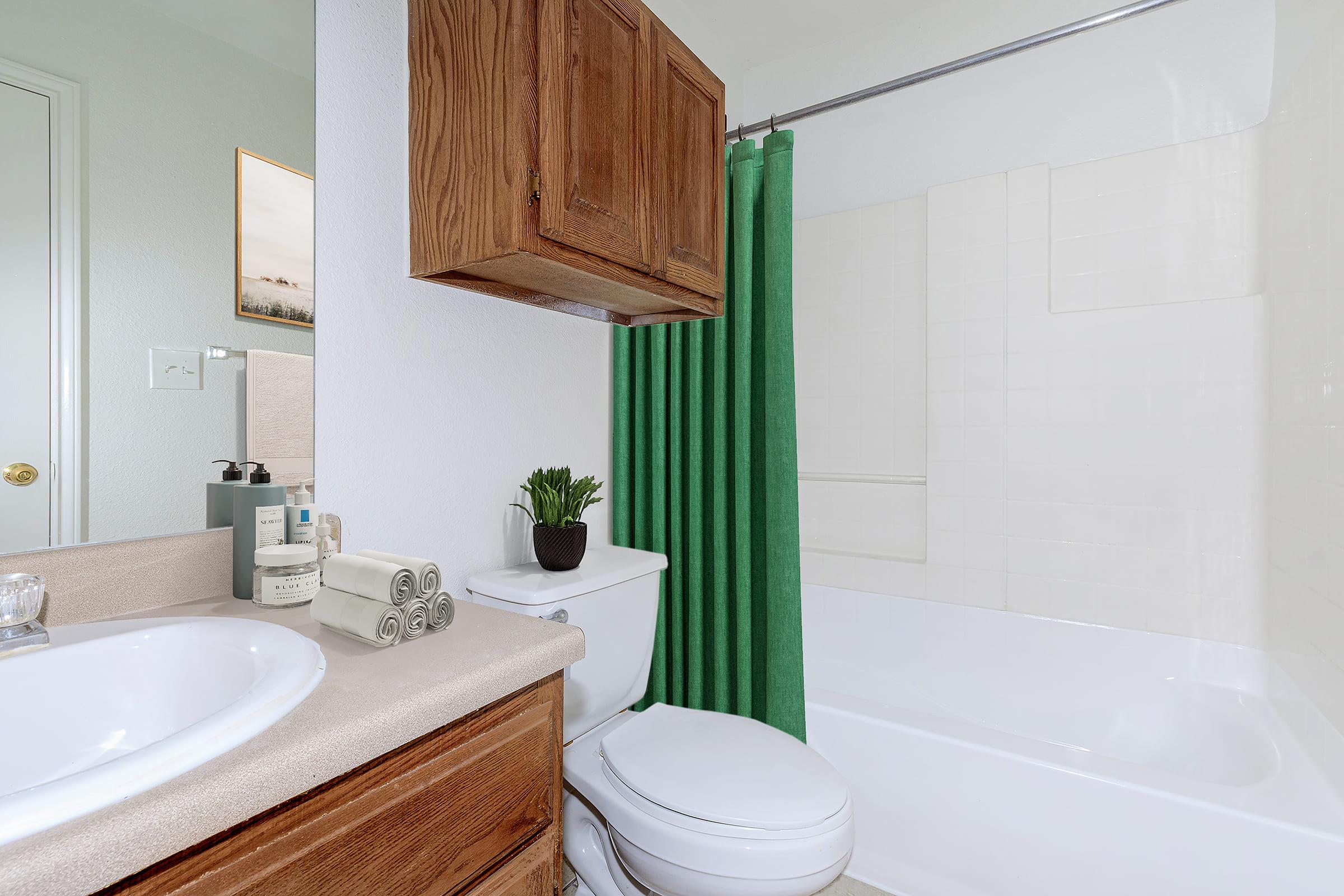 A modern bathroom featuring a white bathtub with a shower curtain, beige countertop with toiletries, and a small plant. Wooden cabinetry is above the toilet, complemented by neatly arranged towels. Natural light illuminates the space through a small window.