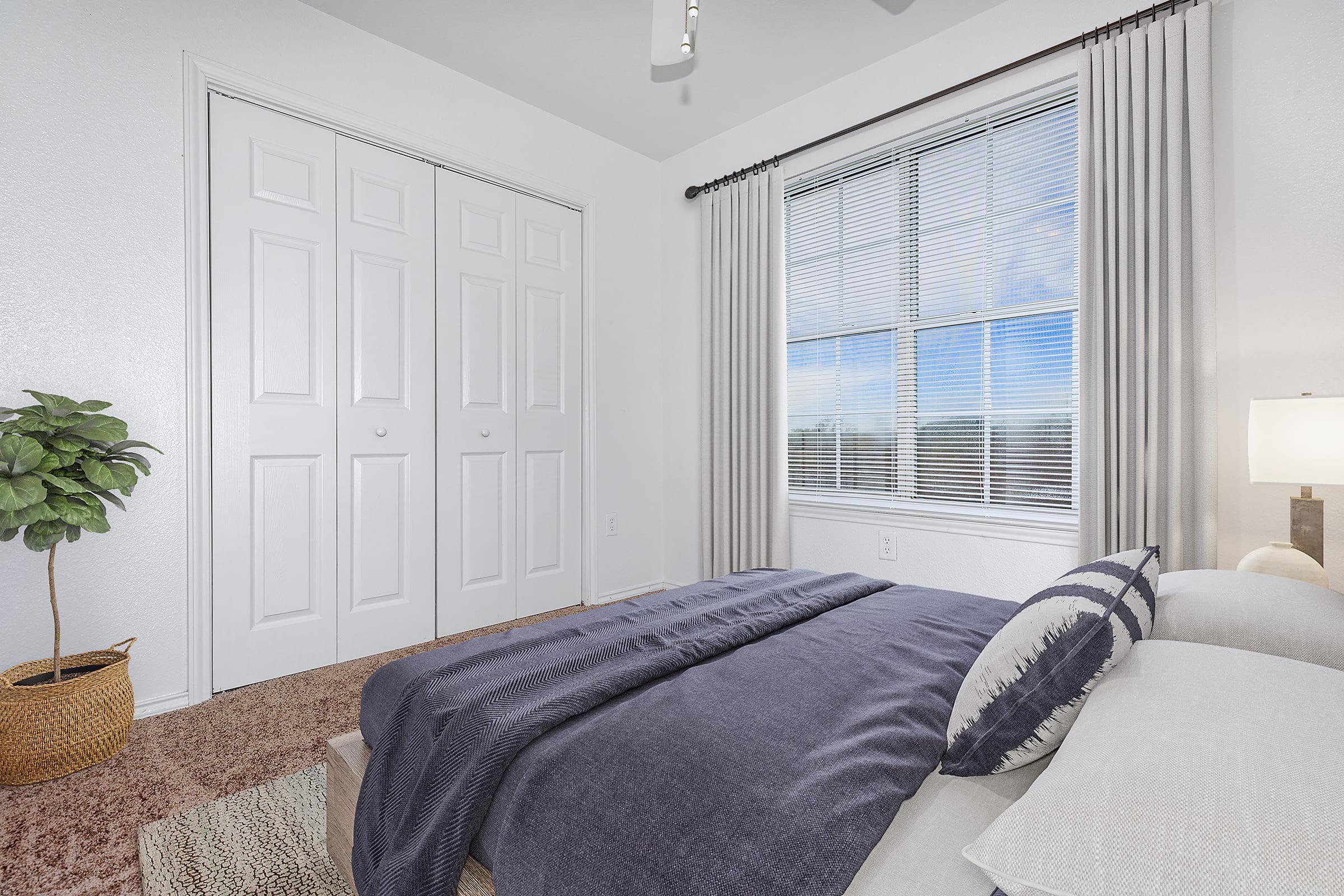 A cozy bedroom featuring a neatly made bed with a navy blue blanket, decorative pillows, and a bedside lamp. The room has light-colored walls, a large window with sheer curtains allowing natural light, and a potted plant in a woven basket. There are dual closet doors visible in the background.