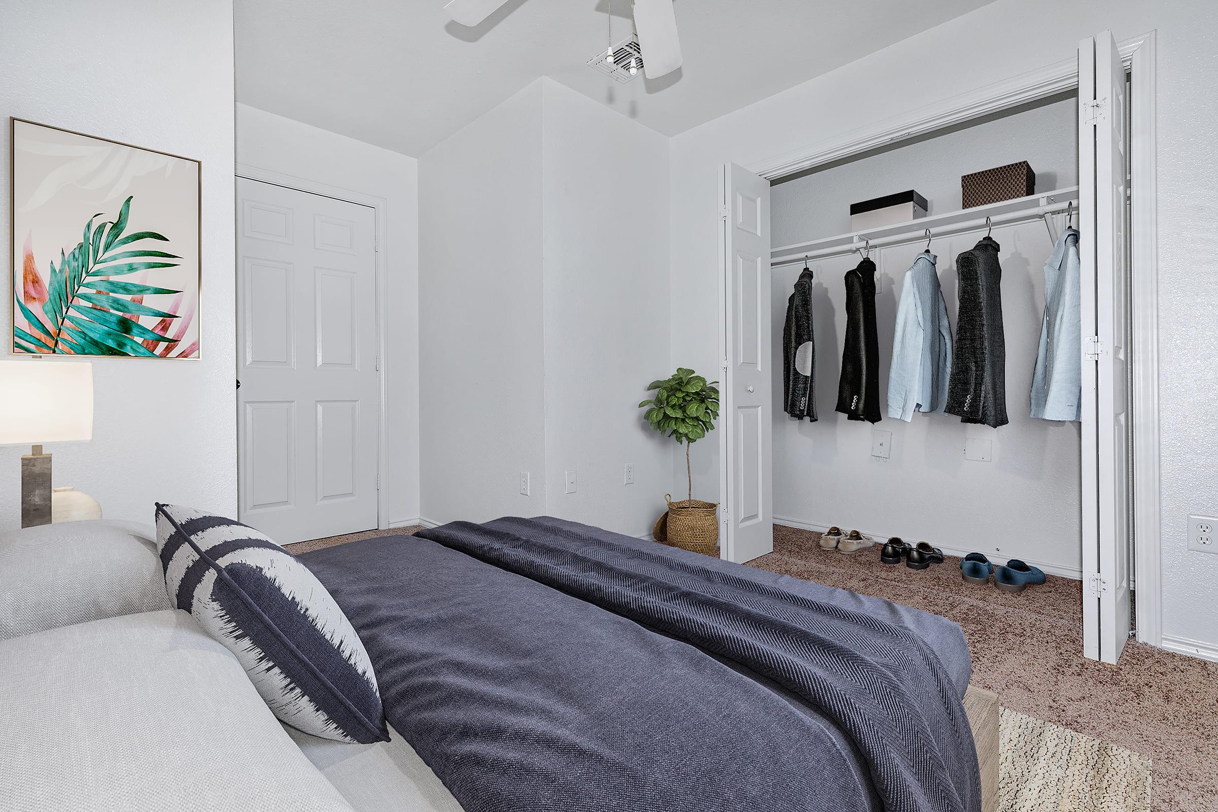A cozy bedroom featuring a bed with a dark blanket, a bedside lamp, and a framed plant artwork on the wall. A closet with hanging clothes is visible, and a small potted plant adds a touch of greenery. The room has neutral-colored walls and carpet, creating a warm and inviting atmosphere.