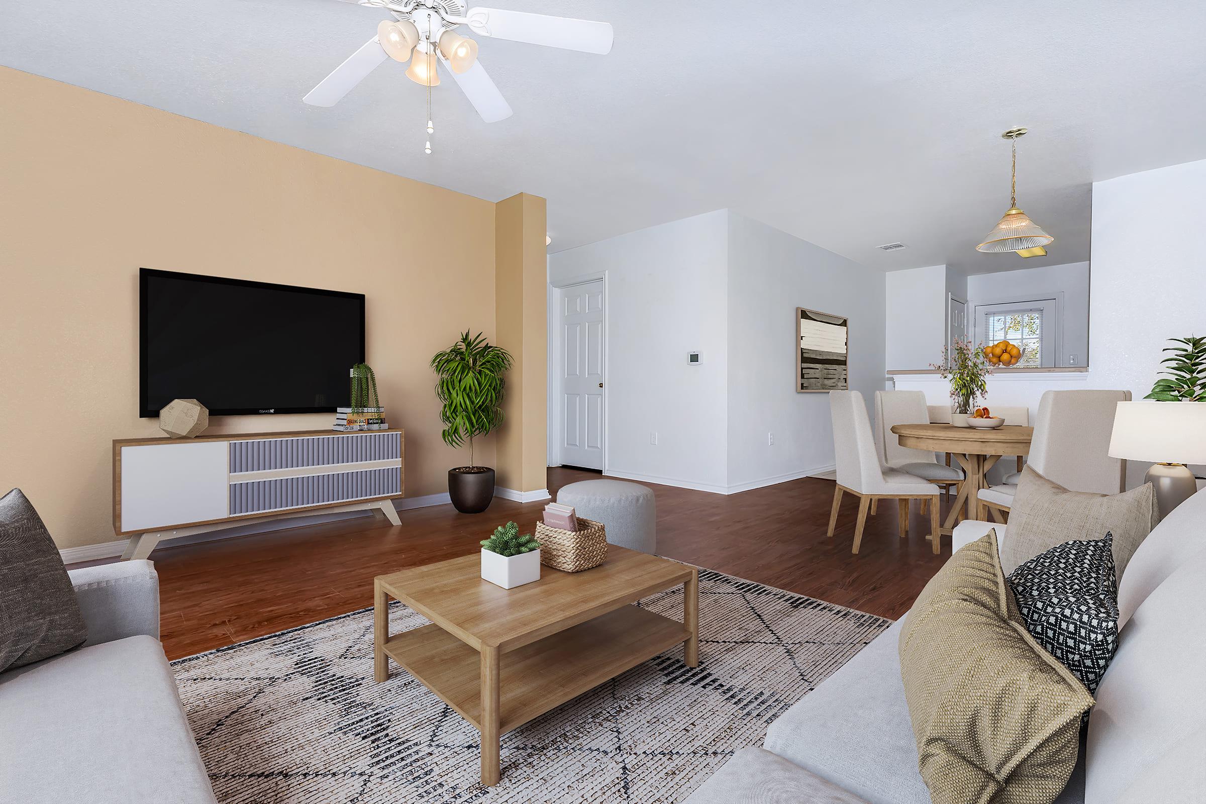 Modern living room featuring a cozy seating area with light gray sofas and a wooden coffee table. A flat-screen TV is mounted on the wall, and there is a decorative plant on the table. The space is illuminated by a ceiling fan and has a warm, neutral color palette, with a dining area visible in the background.